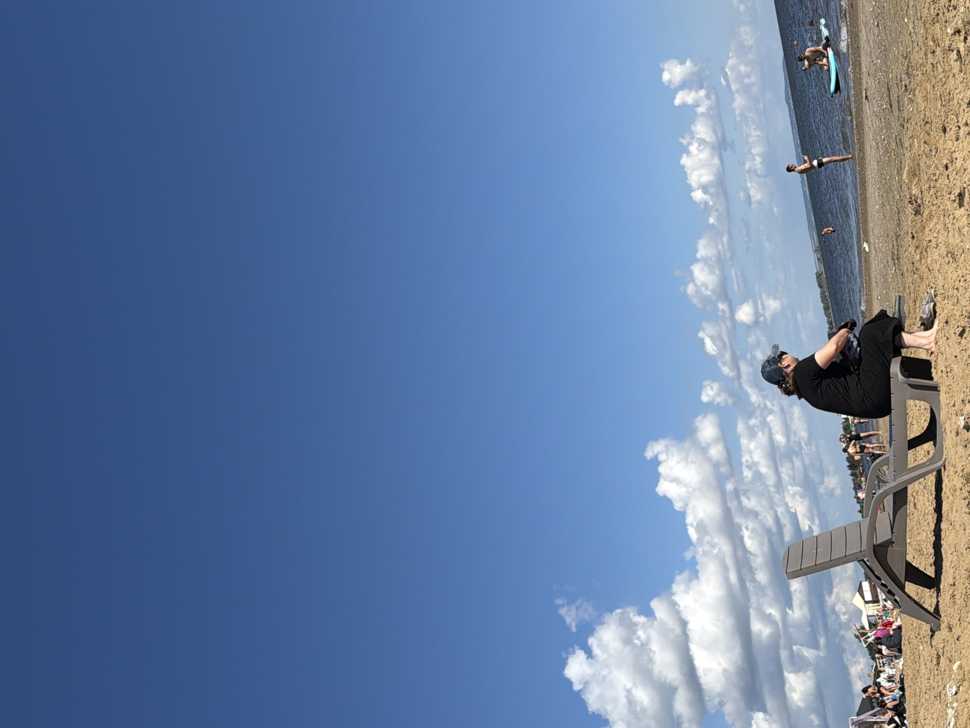 The image depicts a sunny day at a beach with a clear blue sky and scattered white clouds. In the foreground, a person is seated on a gray lounge chair on the sand, facing away from the camera. The person is wearing a dark t-shirt, dark pants, and a blue baseball cap. Their feet are bare, and a pair of sandals are visible near the chair.
In the background, the water of a lake or sea stretches to the horizon. Several people are visible near the water. Some are standing, while others are in or on the water. One person is standing on the shore, another is in the water, and two individuals are on a paddleboard, with one of them appearing to be falling or having just fallen off. Further down the beach, there are more people, some with tents or beach umbrellas. The beach is made of sand. The overall scene conveys a relaxed atmosphere of a summer day at the beach.