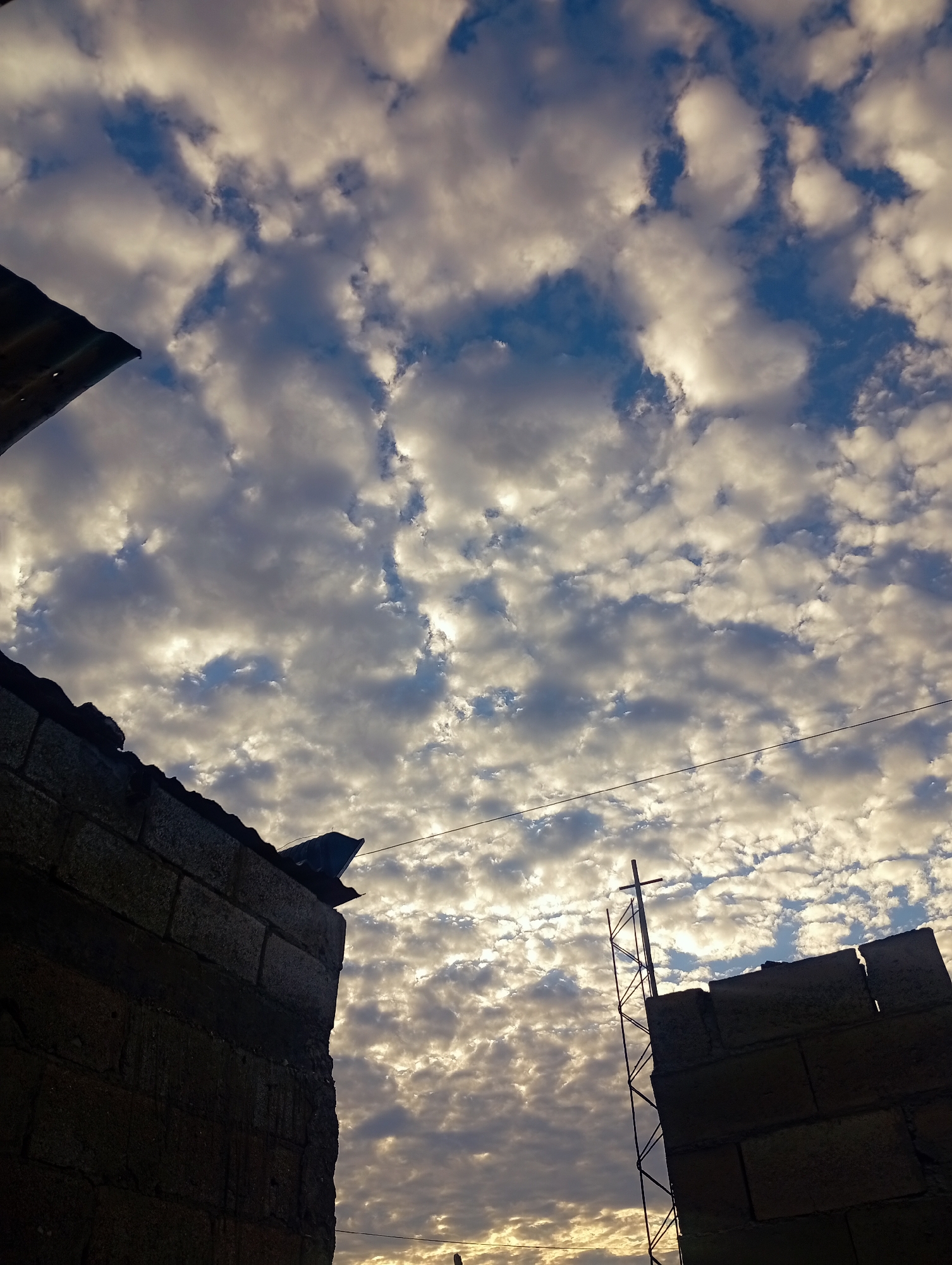 The image shows a low-angle view of a sky filled with altocumulus clouds, punctuated by patches of blue sky. Two unfinished brick structures frame the scene. The structure on the left has a corrugated metal roof section. A metal cross is visible atop the structure on the right. A power line runs horizontally across the image. The location is Altagracia, Venezuela.