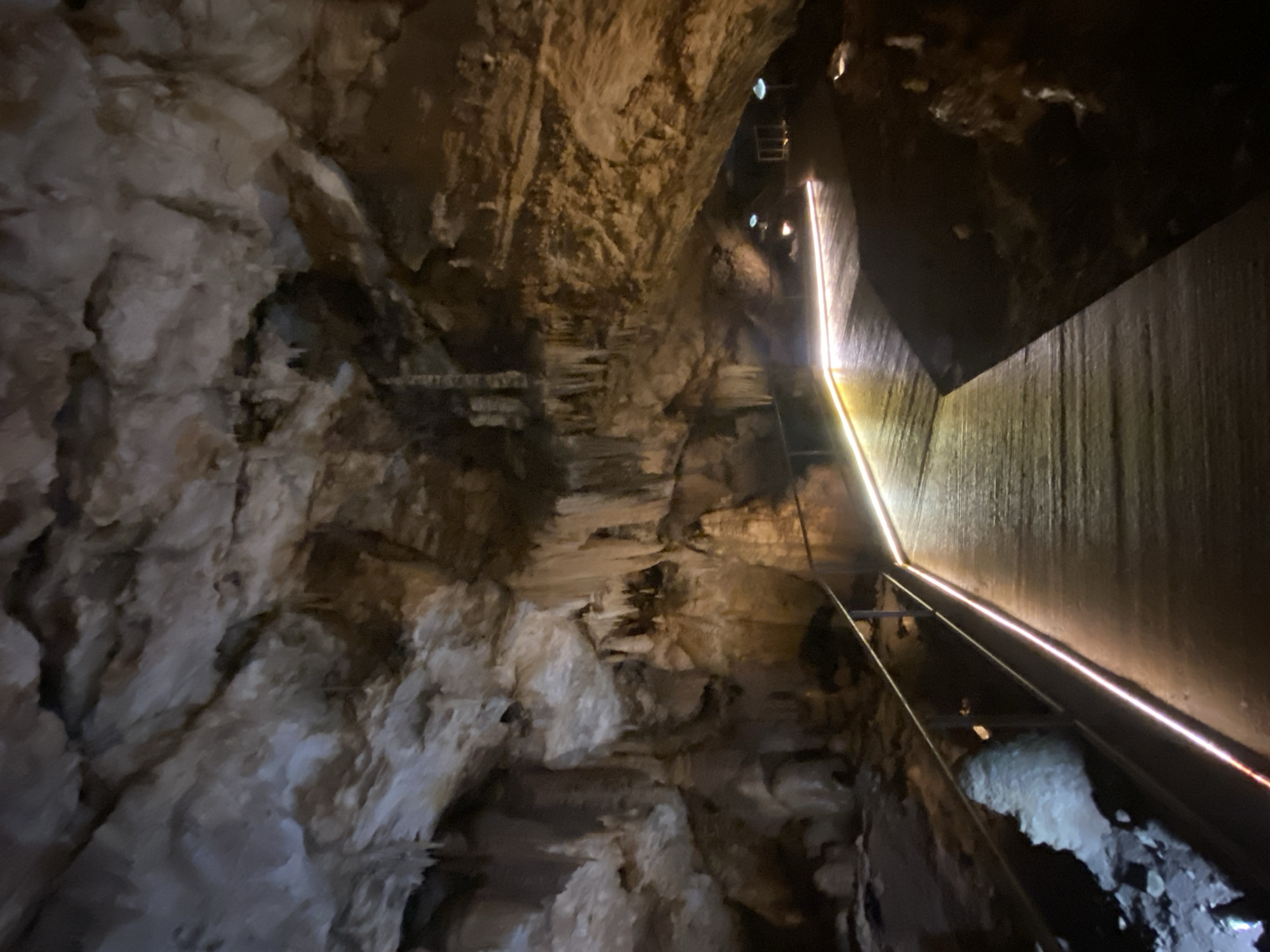 The image shows the interior of Lipa Cave in Lipa Dobrska, Montenegro. A pathway with handrails winds through a cavern. The cave walls are textured and illuminated. Light fixtures are visible in the distance along the pathway. The cave appears to be a tourist destination.