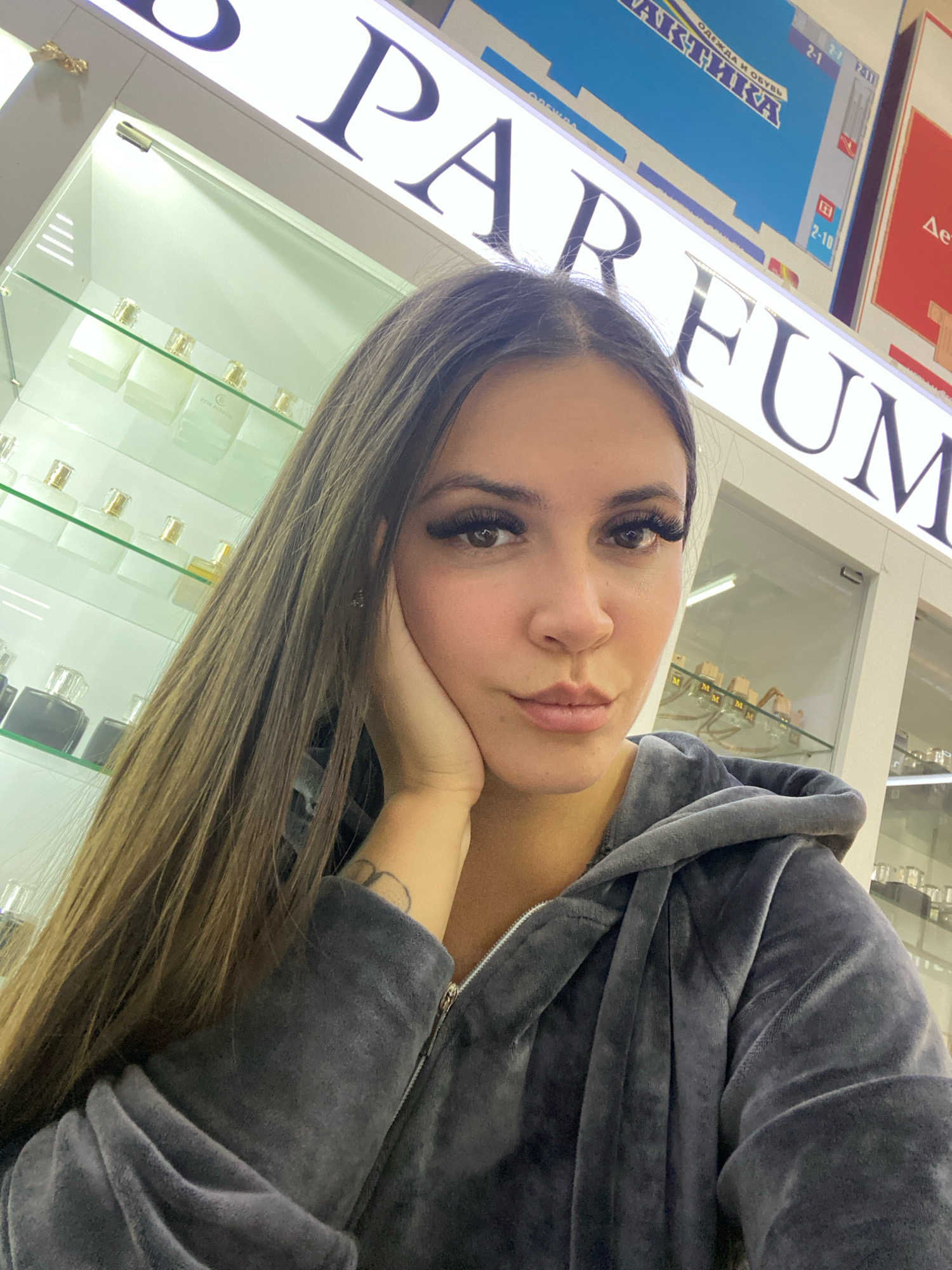 A young woman with long brown hair, wearing a grey velour hooded jacket, takes a selfie in what appears to be a perfume or cosmetics store. She is leaning her head on her hand, with her face partially obscured by her hair. Behind her, shelves are stocked with various perfume bottles. A large sign above the shelves reads "PARFUM" in prominent letters. The store appears to be indoors, well-lit, with a modern and clean aesthetic.