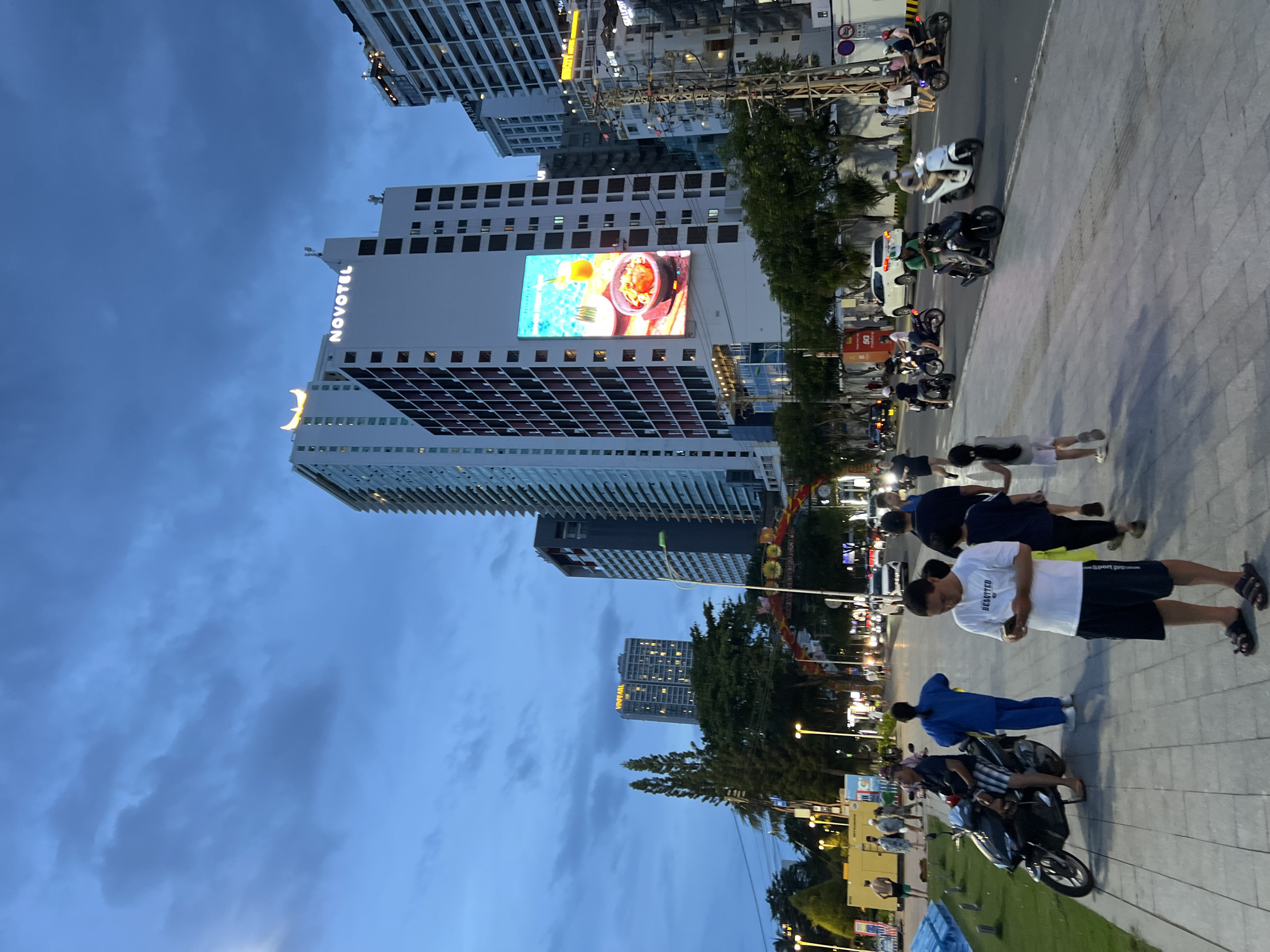 The image shows a waterfront promenade in Nha Trang, Vietnam, at dusk. Tall buildings line the street, including the Novotel with a prominent illuminated sign. A large digital billboard on one of the buildings displays an advertisement for food and drink. People are walking along the promenade and riding motorbikes on the adjacent street. Some individuals are dressed in casual attire, while others appear to be wearing uniforms. The sky is overcast with a few clouds. The overall scene suggests a lively urban environment with ongoing activity.