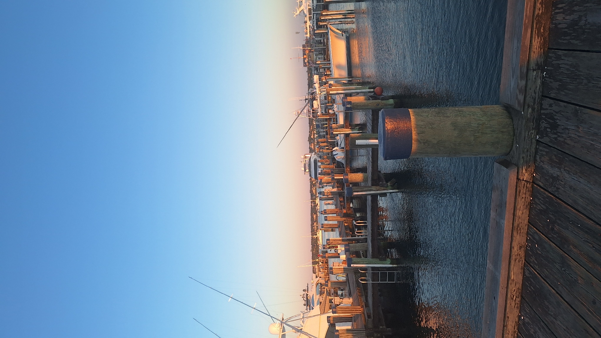 The image depicts a marina in Nantucket, United States of America, at dusk or dawn. The sky is a clear, soft blue, transitioning to a warm, orange hue near the horizon, indicating either sunrise or sunset. The foreground features a wooden dock with weathered planks, leading the eye towards the water.
Dominating the midground is a series of wooden pilings and docks that extend into the water. These structures support several boats of varying sizes. One white boat, prominently positioned on the right, is illuminated by the low-angled sunlight, casting a golden reflection on the dark water. Other boats, including what appear to be larger yachts and fishing vessels, are moored further back, with masts and fishing rods visible. The water is calm and reflects the warm light from the sky and the boats.
In the immediate foreground, a large wooden piling with a dark blue cap stands tall, with its surface reflecting the warm light. Behind this, a ladder is visible attached to a dock, suggesting accessibility to the water or boats. The background reveals a strip of land with buildings and vegetation, characteristic of a coastal town. The overall atmosphere is serene and tranquil, capturing the quiet beauty of a harbor at the edge of day.
