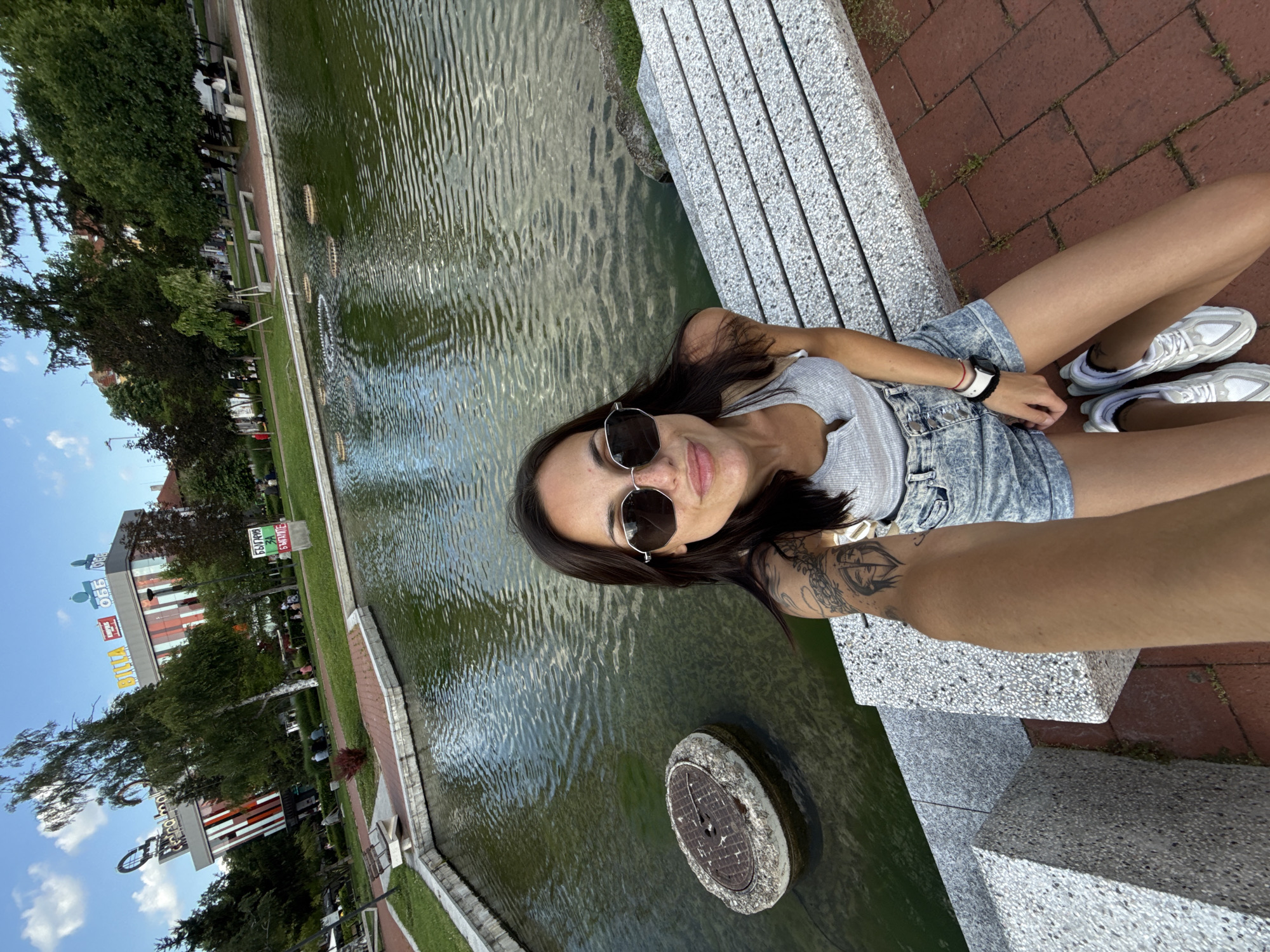 A young woman is depicted in a selfie, taken outdoors in a public park. She is seated on a light grey, speckled stone bench that runs along the edge of a rectangular pond. Her right arm is extended, holding the camera, capturing herself from a high angle. She has dark hair, wears dark hexagonal sunglasses, a light grey ribbed tank top, and acid-wash denim shorts. A white smartwatch is visible on her left wrist, and she has several tattoos on her left arm, including one depicting two intertwined faces. Her white sneakers are visible on the brick-paved ground in front of the bench.
The pond contains greenish-brown water with ripples, reflecting the sky. A corroded, circular metal manhole cover encased in concrete is partially submerged in the water to her left. Further out in the pond, several dark, circular disc-like objects are also partially submerged.
In the background, beyond a grassy area with trees and several people sitting on benches or walking, a large, modern building with a distinctive red and white vertical stripe facade is prominent. Signs for "RetroBoch", "BILLA", and "ОББ" are clearly visible on its upper levels. To the right of this building, a green sign with white Cyrillic text reads "БЪЛГАРИЯ ЗА БЪЛГАРИТЕ" (Bulgaria for Bulgarians). The sky is clear blue with sparse white clouds. This location is identifiable as a public space near the RetroBoch shopping center and the Water Cascade (Kaskada) area in Pleven, Bulgaria.