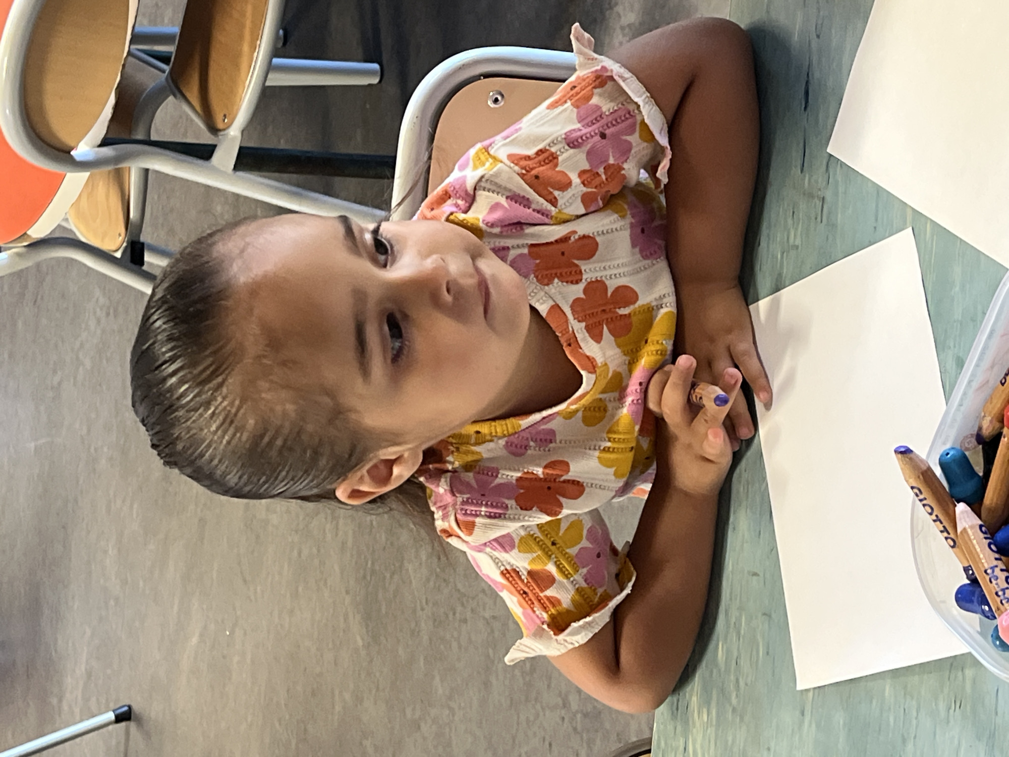 A young girl is sitting at a desk, looking to her left. She wears a floral patterned top. Her hair is dark and appears wet. She is holding a crayon in her right hand. In front of her is a blank sheet of white paper, and a container of crayons sits to the right. There are chairs visible in the background. The location appears to be indoors. There is no information to verify that the location is in the city of La Fouillouse, France.