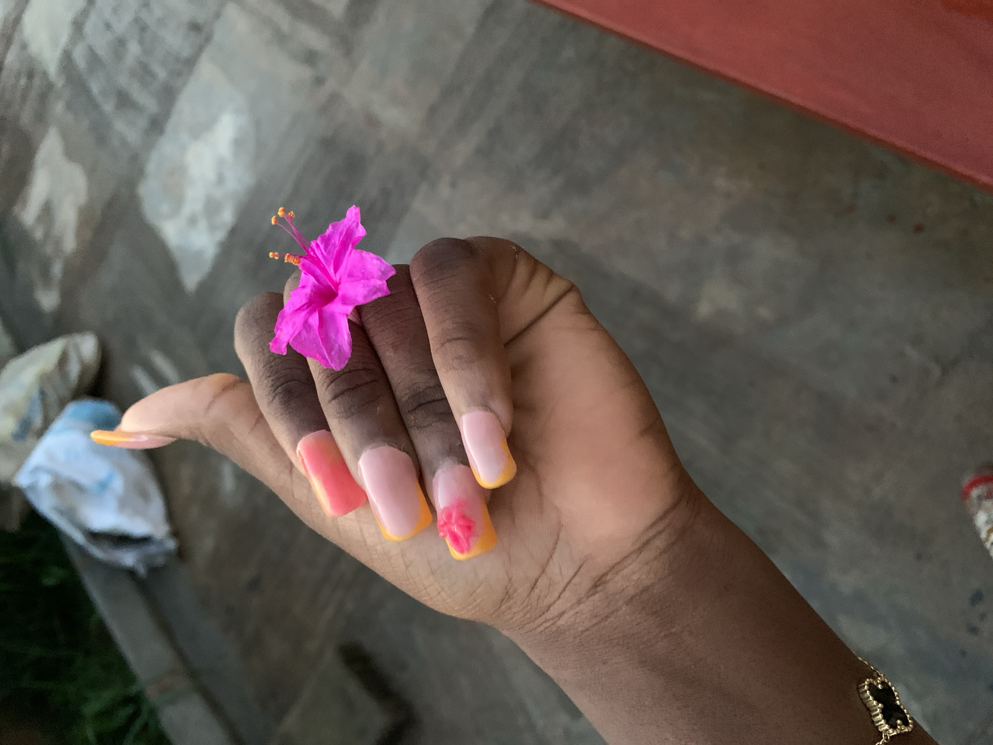 An overhead medium shot shows a person's left hand with manicured nails, a bright pink flower resting on the index finger, and a gold necklace with a black charm on the wrist. The nails are long and square-shaped, with a French manicure design featuring a sheer pink base and a gradient of orange and pink at the tips. The flower appears to be a type of hibiscus, with delicate pink petals and orange stamens. The hand is positioned over a grey, textured surface, possibly a patio or pavement. In the background, out of focus, are some white and brown plastic bags and green foliage. The lighting suggests an outdoor setting, possibly during the day. The image is taken from a slightly elevated perspective.