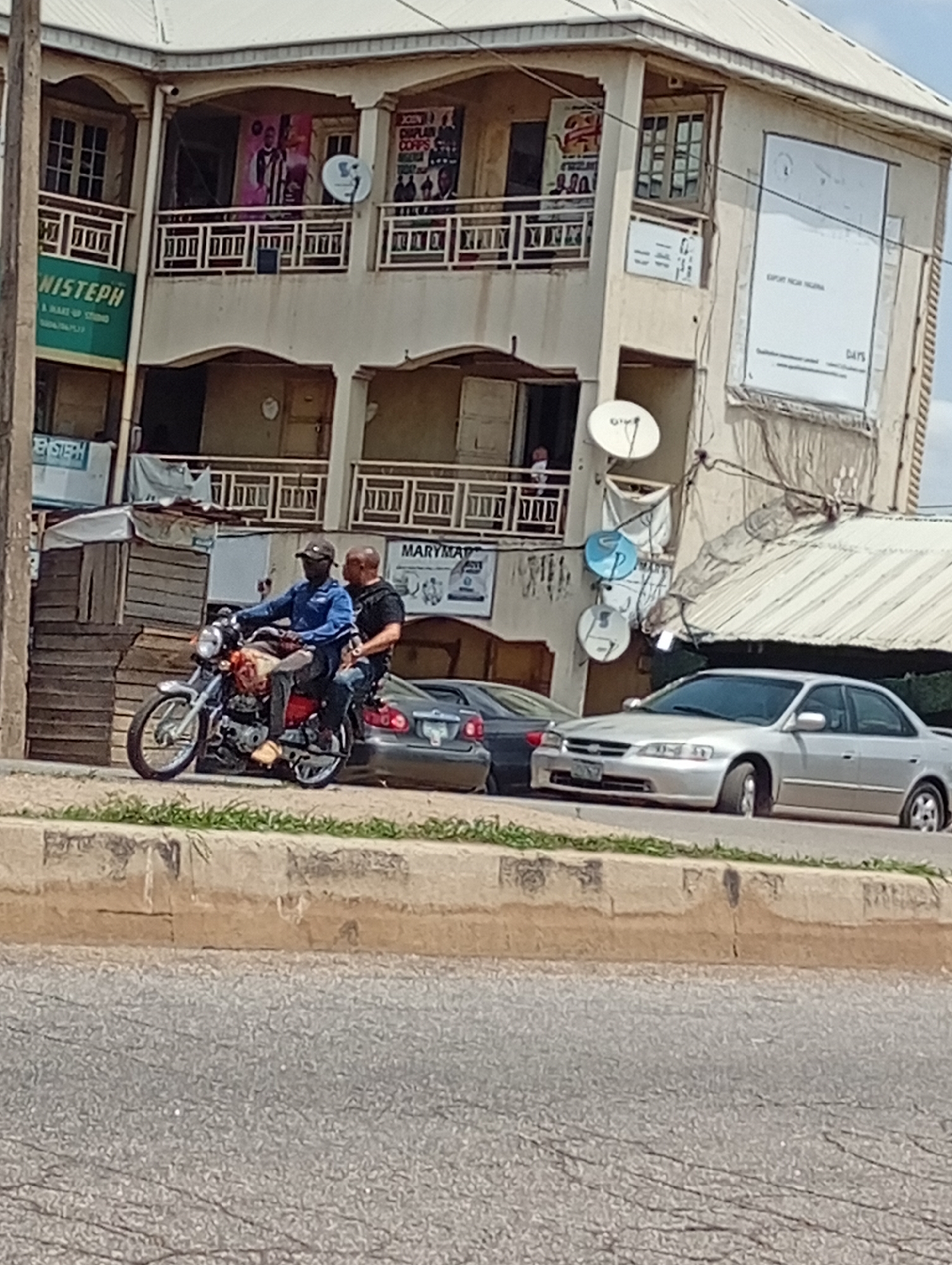 The image captures a street scene in what appears to be an urban setting. A red motorcycle with two male riders is the central subject. The driver is wearing a blue shirt and cap, while the passenger is in a black shirt. Behind them, two cars, one gray and one dark, are parked along the curb.
The backdrop consists of a multi-story building with balconies, displaying various advertisements and signs including "NISTEPH" and "MARYMAI." Satellite dishes are attached to the building's exterior. The street itself is asphalt with a raised curb. The location is in the city of Unknown, Unknown.