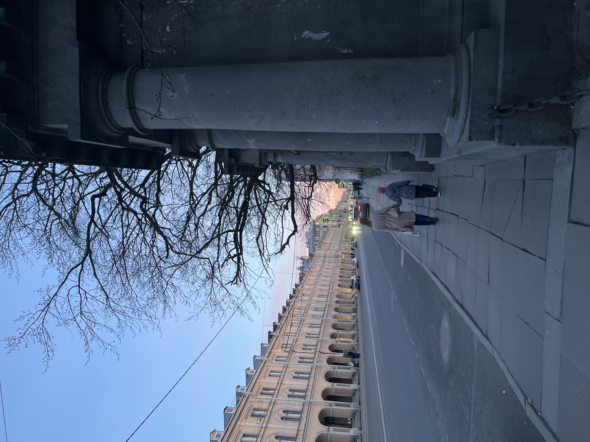 The image captures an urban street scene at what appears to be dusk or dawn, given the subdued light and illuminated streetlights. The perspective is from beneath a large structure, looking down a long street.
In the immediate foreground, a dense network of bare tree branches, devoid of leaves, stretches across the top of the frame against a clear, light blue sky. This suggests either late winter or early spring.
Dominating the midground are imposing, grey stone columns, forming a colonnade that runs parallel to the street on the left side of the image. The columns are thick and sturdy, indicative of classical architecture. Below these columns, on a paved sidewalk, two individuals are walking away from the viewer. The person closer to the camera wears a light-colored long coat over blue pants. Further ahead, another person is dressed in a dark jacket and a distinctive red beanie.
To the right of the columns, a wide, empty street or boulevard stretches into the distance, with a few parked cars visible further down. The streetlights along this road are illuminated, casting a warm glow.
On the far right, lining the street, is a long, multi-story building with a light-colored facade, featuring numerous arched windows or niches. The building's architectural style appears grand and historical. The sky behind this building transitions from light blue to a faint pinkish-orange hue on the horizon, reinforcing the sense of early morning or late evening.
There is no discernible text visible in the image. The overall atmosphere is quiet and calm.