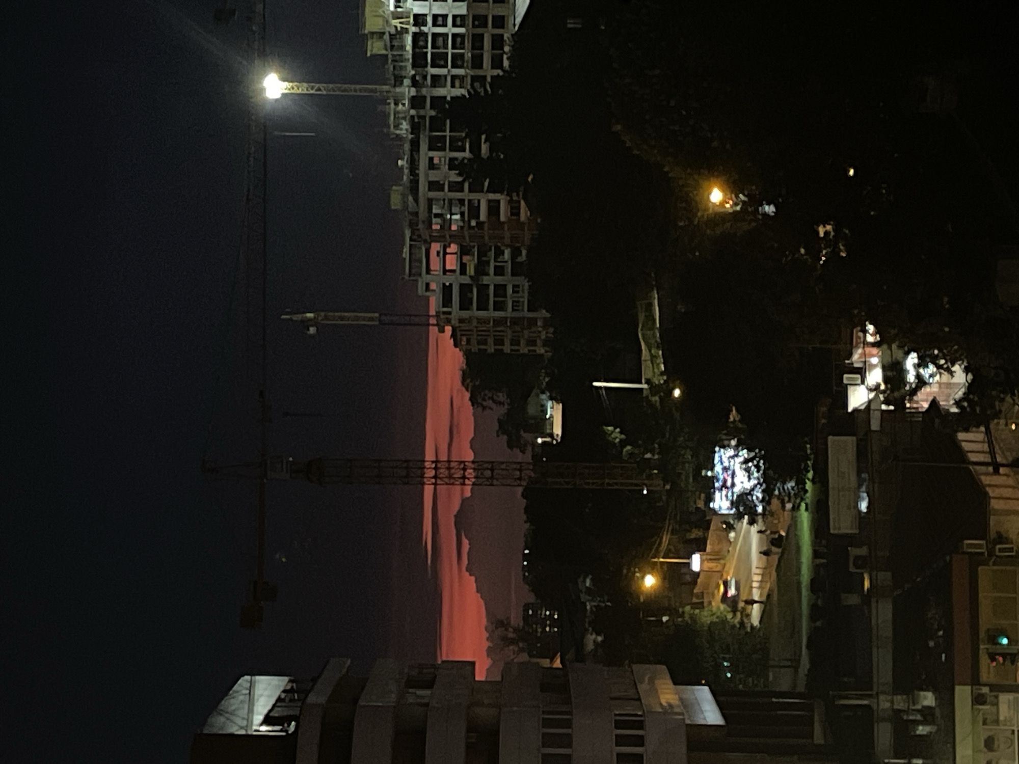 The image captures a night scene in Belgrade, Serbia, featuring construction sites with cranes against a backdrop of a colorful sunset sky. The foreground shows rooftops of buildings. Streetlights illuminate the cityscape, and trees are silhouetted. The cranes and unfinished building indicate ongoing construction activity.