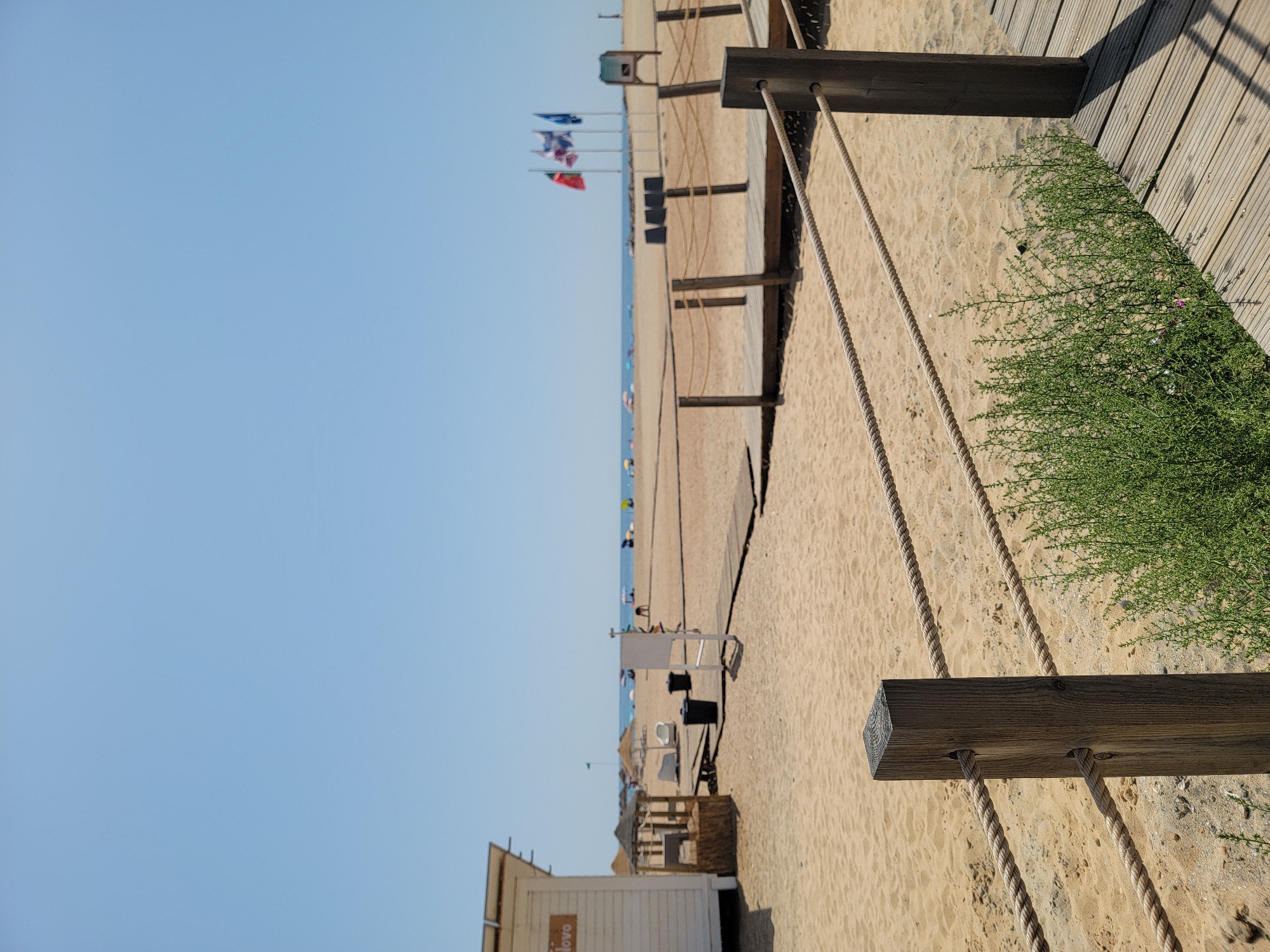 The image depicts a sandy beach in Quarteira, Portugal, on a clear, sunny day. In the foreground, a wooden walkway with a rope railing is visible, bordered by green shrubbery. The walkway leads towards the beach, where several black trash bins and some beach chairs are scattered. A small white wooden structure with the word "Novo" visible on its side is located to the left of the frame. In the distance, the sea is calm under a bright blue sky. Several flags, including what appears to be the Portuguese flag, are flying on poles further down the beach, near a small structure. A few beach umbrellas and people are sparsely distributed across the sand, suggesting a relatively uncrowded beach environment. The scene conveys a typical summer day at a coastal resort.
