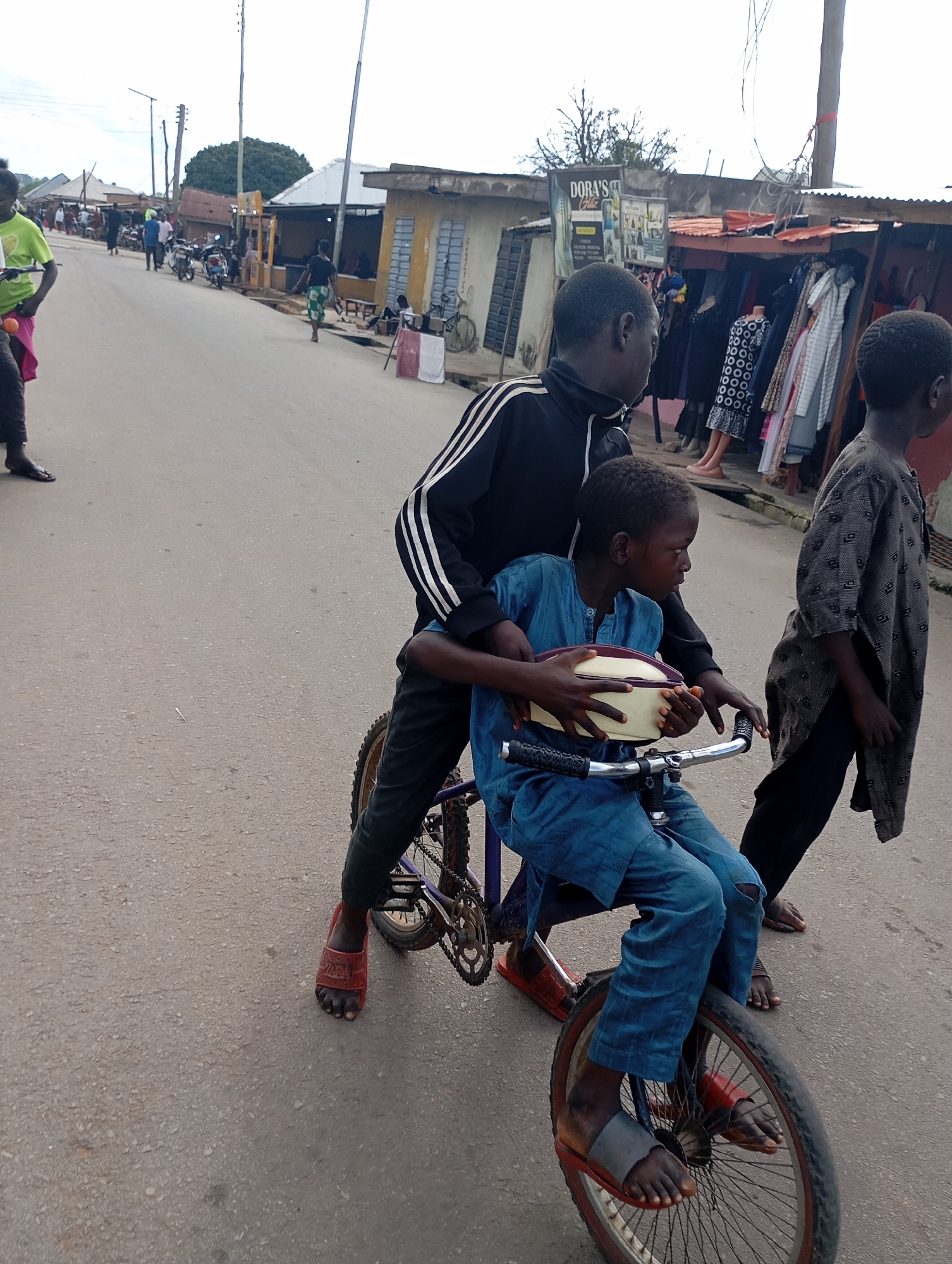 The image depicts a street scene in Shendam, Nigeria, with two boys on a bicycle in the foreground. The boy in front, wearing a blue outfit, is seated on the bicycle and holding a beige and white oval object. The boy behind him, wearing a black tracksuit with white stripes, has his arms around the front boy and appears to be assisting him. Both boys are wearing sandals. Behind them, a third boy in a patterned gray garment walks along the street. The environment includes a paved road lined with shops on either side. Various clothing items are displayed for sale outside one of the shops. Several motorcycles are parked along the street, and other individuals can be seen in the background. The overall atmosphere suggests a bustling market or commercial area.