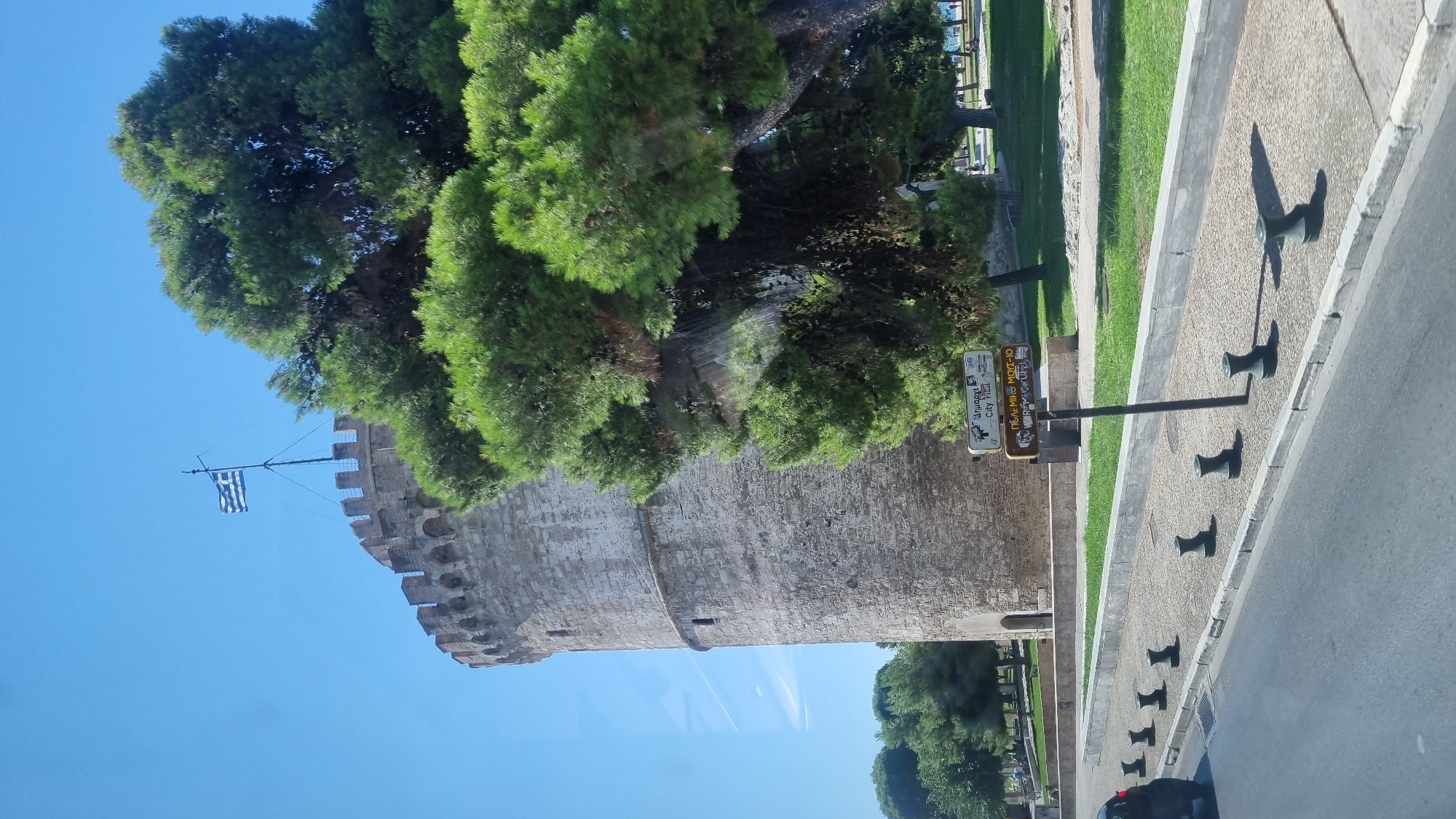The image shows the White Tower in Thessaloniki, Greece. The tower is a prominent landmark of the city, located in the Thessaloniki Municipal Unit. The tower is cylindrical and made of stone, with battlements at the top. A Greek flag flies from a flagpole on top of the tower. A large pine tree with dense green foliage is in the foreground, partially obscuring the tower. In the middle ground, a sidewalk is lined with black bollards. Several signs on a post indicate directions to various locations, including "City View." A road is visible in the foreground with the rear of a black car on the left side of the frame. The sky is clear and blue.
