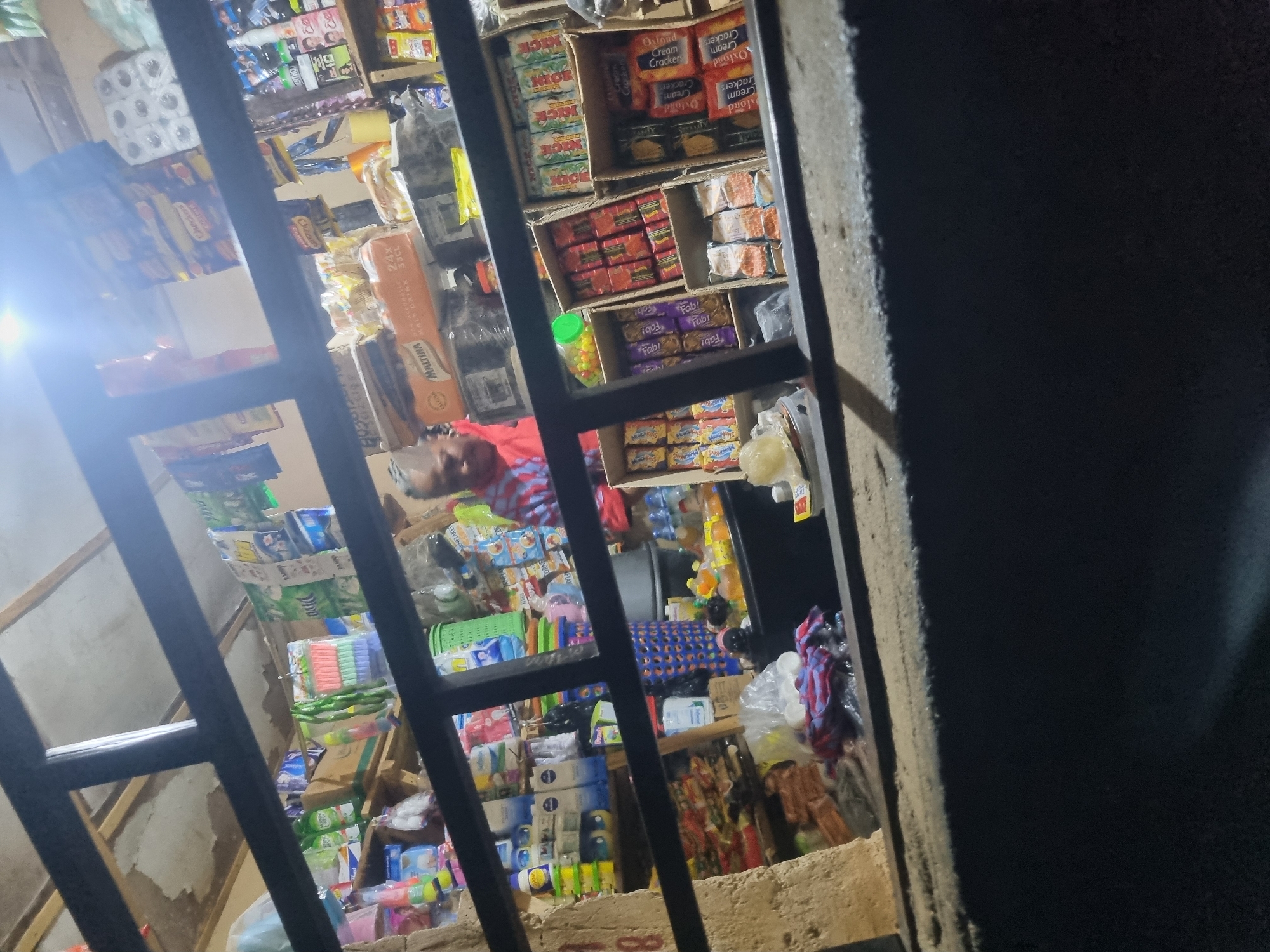 The image depicts a small convenience store, likely a kiosk or a corner shop, in Jos, Nigeria. The store is filled with various retail goods, including packaged food items, toiletries, and cleaning supplies, neatly arranged on shelves and in display boxes. The lighting suggests it is nighttime or dusk, with artificial light illuminating the interior. A person, possibly the shopkeeper, is visible in the background, wearing a red and blue striped shirt, and appears to be attending to the store. The store is partially obscured by a dark, horizontal metal grate or bars, suggesting the viewer is looking into the store from the outside. The overall scene conveys a sense of a typical urban retail environment in Nigeria.