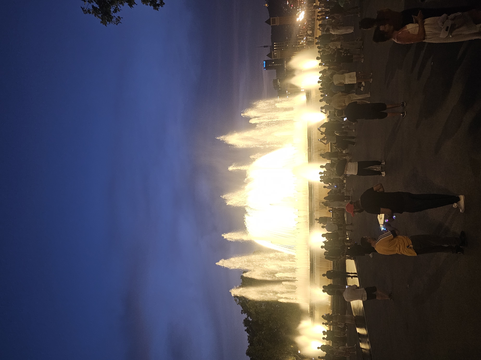 The image depicts a nighttime scene at the Magic Fountain of Montjuïc in Barcelona, Spain. A large, illuminated fountain is actively spraying water, creating a dramatic effect against the dark blue twilight sky. Numerous people are gathered in the foreground and middle ground, observing the fountain. Many are holding up smartphones, likely to take pictures or videos of the spectacle. The crowd is diverse, with individuals and groups of varying ages. The environment appears to be an open plaza with trees visible in the background, bordering the fountain area. Lights from buildings can be seen in the distance, suggesting an urban setting. The overall atmosphere is one of public enjoyment and appreciation of a nighttime attraction.