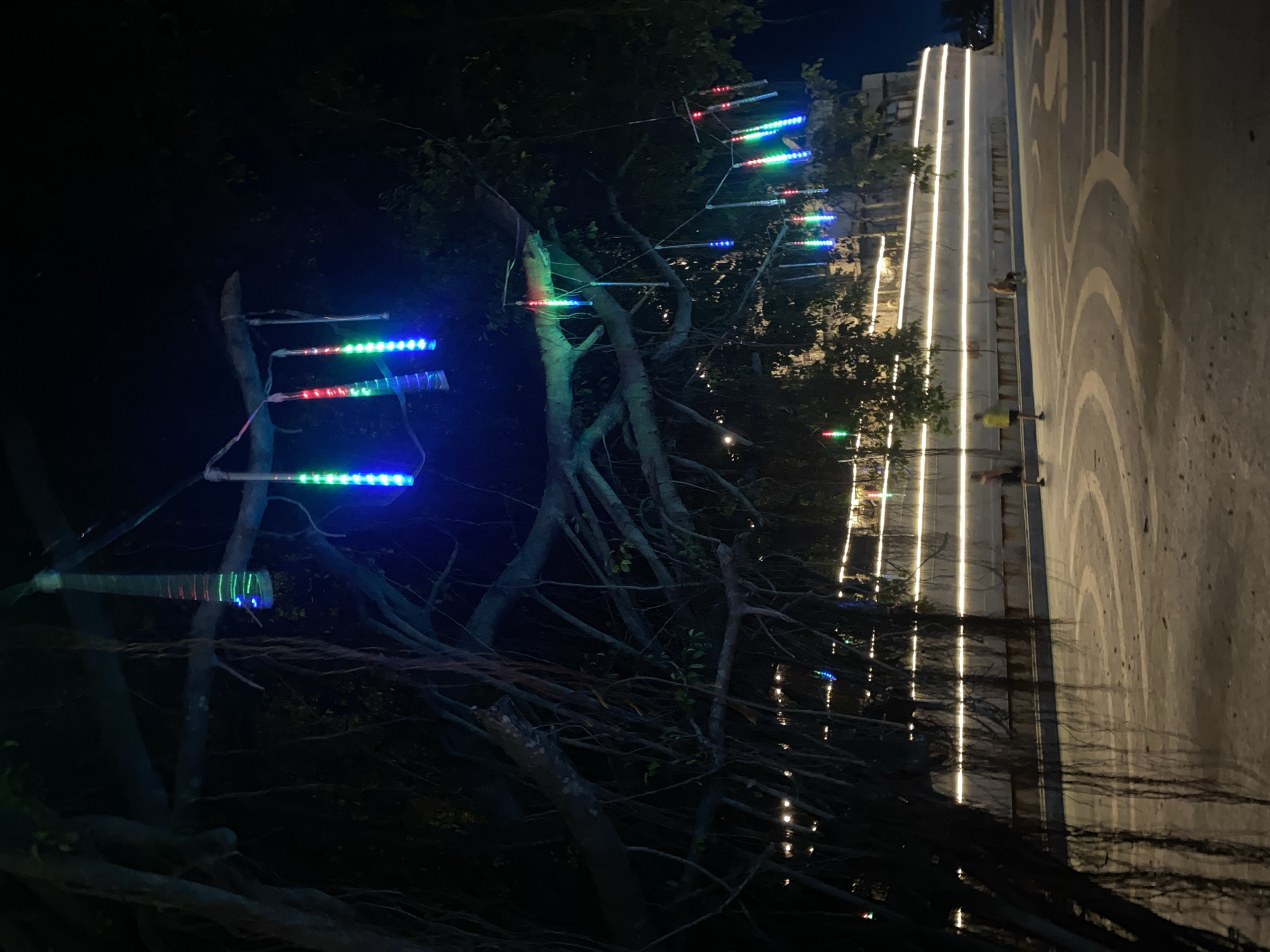 The image shows an outdoor scene at night in Baan Don Sai, Thailand. The primary focus is on trees decorated with LED lights, some displaying red, green, and blue colors. These lights are affixed to branches with what appears to be clear tape. In the background, a path or walkway is illuminated by vertical white light strips. Several indistinct figures are visible on the walkway. The walkway surface is marked with a pattern of concentric circles. The overall environment is dark, with lighting emphasizing the decorated trees and pathway.