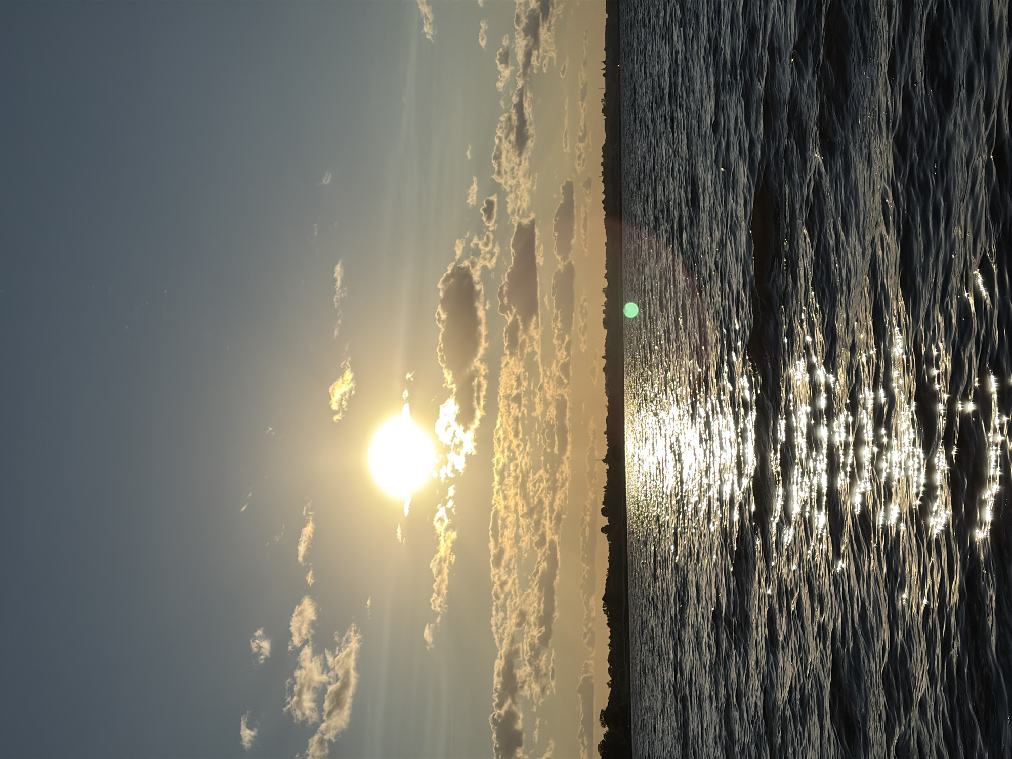 This is a photograph of a body of water, likely a river or lake, with a sunset visible in the background. The sun is a bright orb in the sky, partially obscured by scattered clouds which are illuminated by the setting sun, casting golden hues. A distant shoreline with trees and some structures is visible. The water's surface is rippled, reflecting the sunlight in a shimmering path. The overall scene evokes a sense of tranquility and the beauty of nature. The location provided, Vidogoshchi, Russia, suggests this is a natural landscape, possibly a popular spot for observing sunsets.