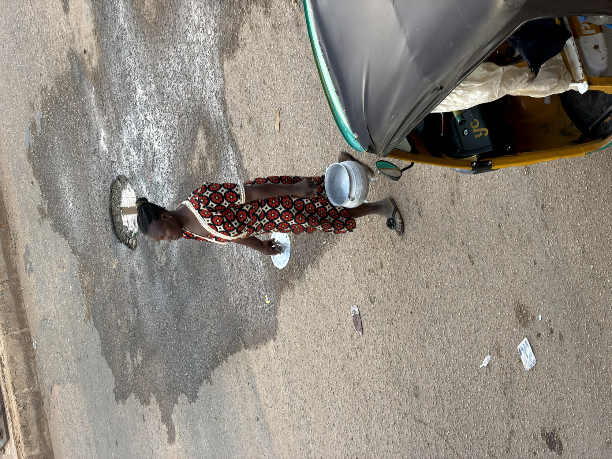 In Bukuru, Nigeria, a woman walks on a wet, asphalt road, carrying a metal pot and a round lid. She is wearing a patterned dress with red, black, and white circles and flowers, and flip-flops. To the right, a portion of a yellow and black auto-rickshaw is visible, with a dark canvas roof and a beige interior lining. The road surface is textured and appears to have several puddles and a large pothole. Scattered debris, including a piece of crumpled paper and what looks like a plastic wrapper, is present on the ground.