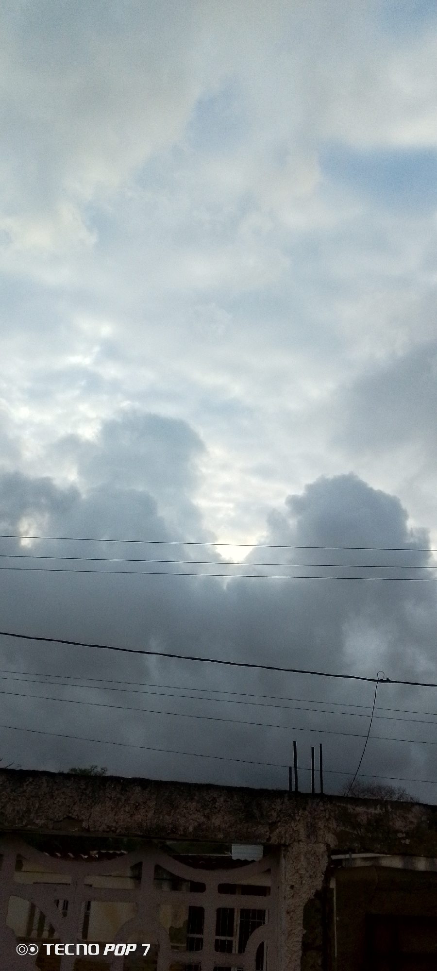 This is a vertical, outdoor photograph of a cloudy sky. The sky takes up the majority of the frame, with lighter clouds towards the top and darker, more textured clouds lower down. A few power lines are visible strung horizontally across the middle of the image. Below the power lines, the top of a wall or fence is visible, with some rebar protruding from the top. The overall impression is one of an overcast or stormy day. The image has a watermark in the bottom left corner that reads "TECNO POP 7". The lighting suggests it could be daytime, possibly late afternoon or early morning, given the muted light. The presence of power lines and a wall hints at an urban or semi-urban setting.