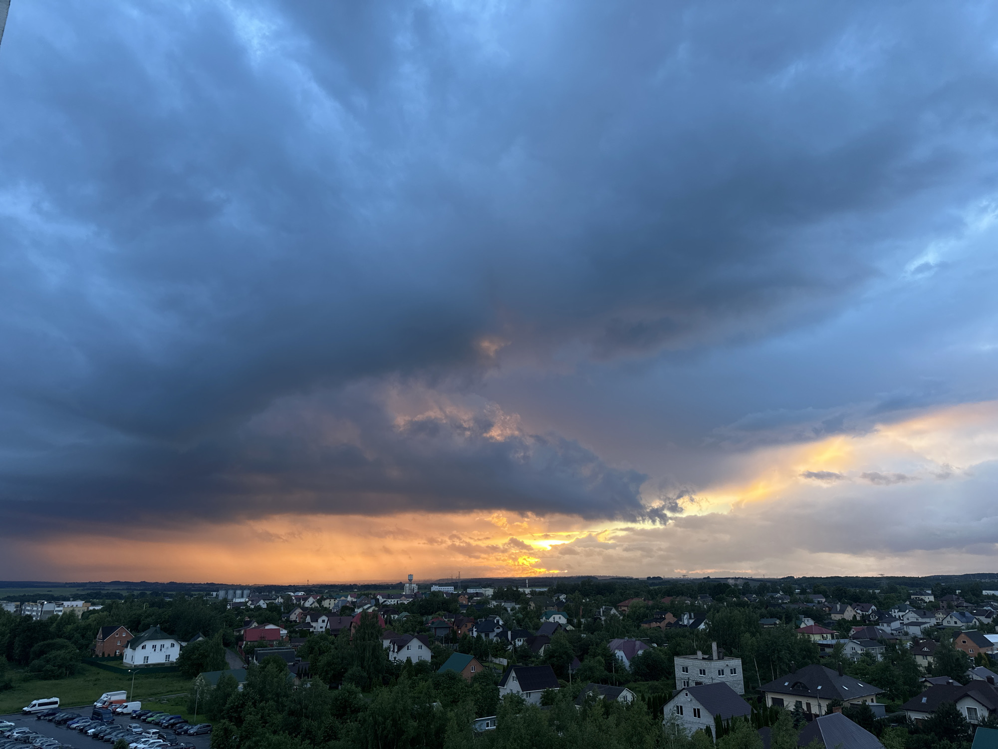 This is an aerial view of a residential area within the Shchomyslitsky rural council in Belarus. The scene is dominated by a dramatic sky filled with dark, heavy clouds, indicative of an approaching storm, contrasted with streaks of vibrant orange and yellow sunset light breaking through. Below the sky, a dense landscape of houses with varied roof colors and styles is visible, interspersed with lush green trees. A parking lot filled with cars is present in the lower left corner, and a water tower can be seen in the distance on the left side of the image.