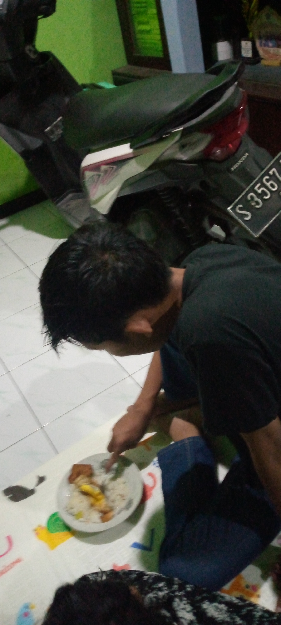 A medium shot captures a person eating a meal on the floor in what appears to be a home in Sumberwono, Indonesia. The person, whose head and torso are visible, is wearing a black t-shirt and blue jeans and is seated on a colorful mat. They are using a spoon to eat from a plate of rice and what appears to be fried food. To the right of the person is a motorcycle, its license plate clearly visible as "S 3567". The background shows a green wall and a shelf with decorative items, suggesting an indoor environment. The lighting is dim, casting shadows on the floor.