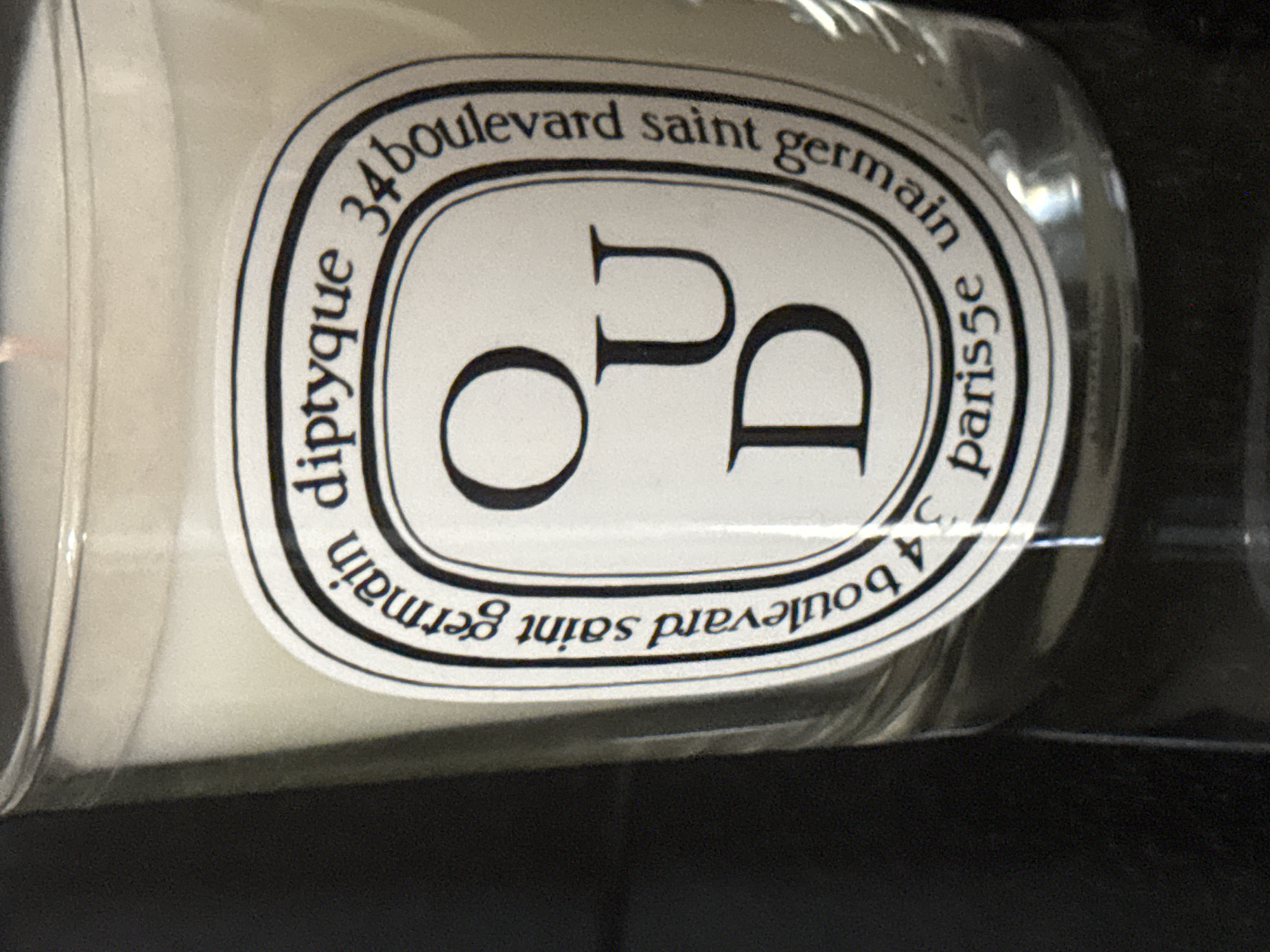 This media file is a close-up, angled shot of a white candle in a clear glass jar. The candle has a label with text printed around an oval shape. The text on the label appears to be in French and English.
The top arc of the oval reads "34 boulevard saint germain". Inside the oval, large letters spell out "OU D". Below that, the arc reads "4 boulevard saint germain" and "paris5e". The word "diptyque" is printed vertically along the left side of the oval, and a small "C." is visible on the right side. The candle itself appears to be made of a solid white material, possibly wax. The lighting is artificial, and the background is dark, suggesting the photograph was taken indoors in a low-light environment. No people or specific activities are depicted. The overall impression is that of a product shot, focusing on the branding and design of the candle.