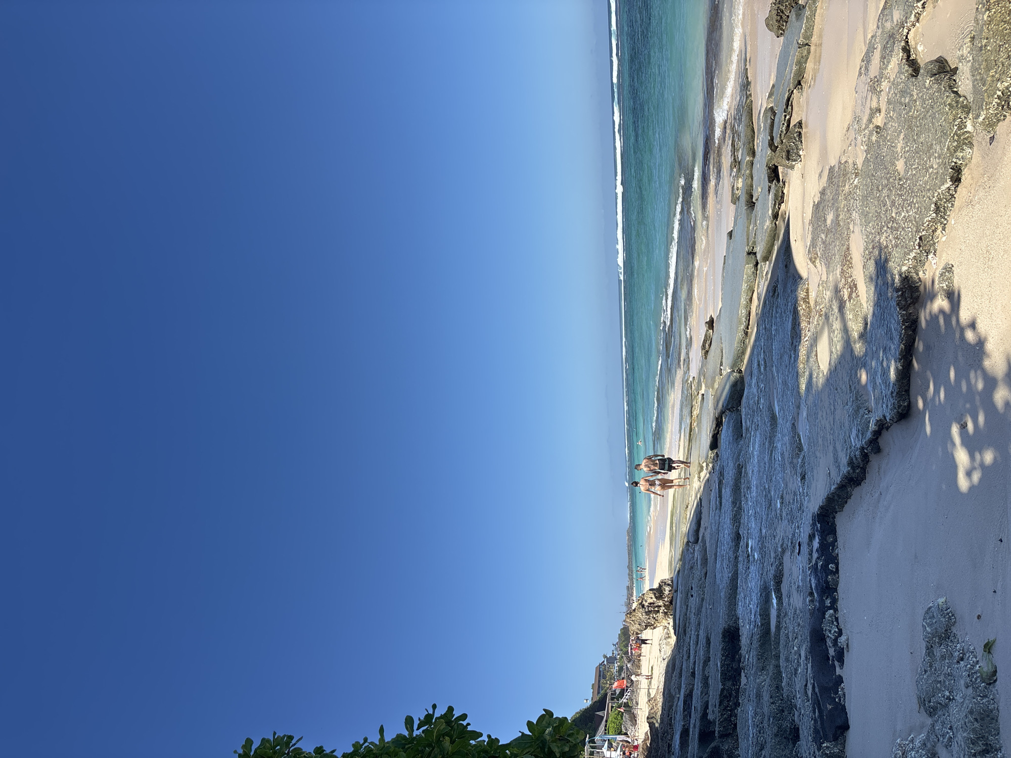 The image shows a beach scene in Kuta Selatan, Indonesia. The sky is clear blue, taking up most of the image. The ocean is visible as a narrow strip with turquoise water and small waves. A sandy beach is present, bordered by rocky formations. Several people are walking on the beach. The foreground includes rocky terrain, possibly a cliffside. Green foliage is visible at the bottom left of the image.