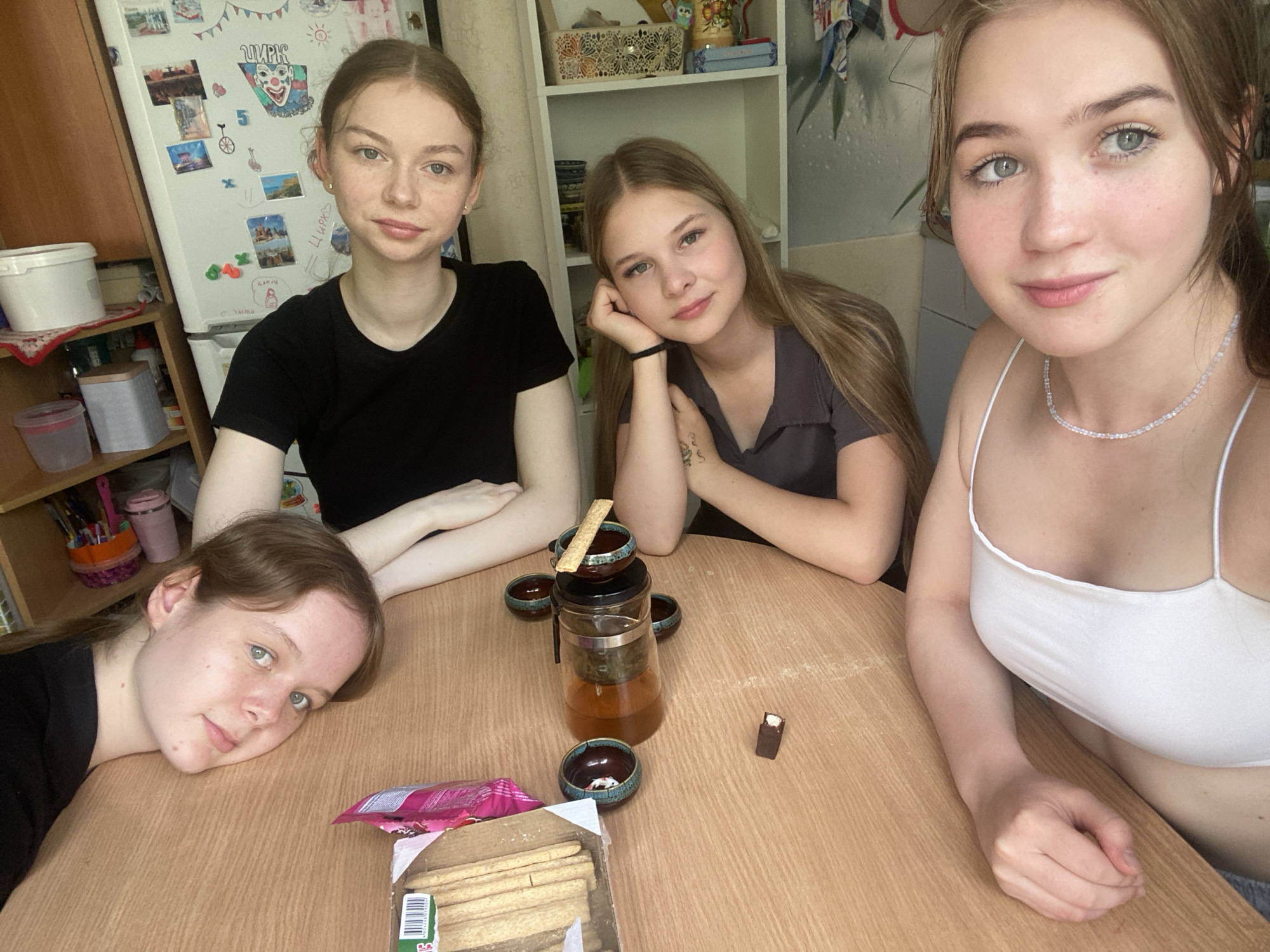 Four young women are gathered around a wooden table, possibly in a kitchen or dining area, in Chaikovsky, Russia. On the table are a glass teapot with amber liquid, several small ceramic cups, a box of cookies, and a single piece of chocolate. The woman on the far left rests her head on the table. The woman next to her, wearing a black t-shirt, is looking towards the camera. The third woman, with long blonde hair, leans on her hand, looking at the camera with a slight smile. The fourth woman, in a white tank top, is positioned closer to the camera and is also looking at the viewer. In the background, a refrigerator is visible, decorated with various magnets and pictures. Shelving units filled with items are also present. The overall scene suggests a casual gathering over tea and snacks.