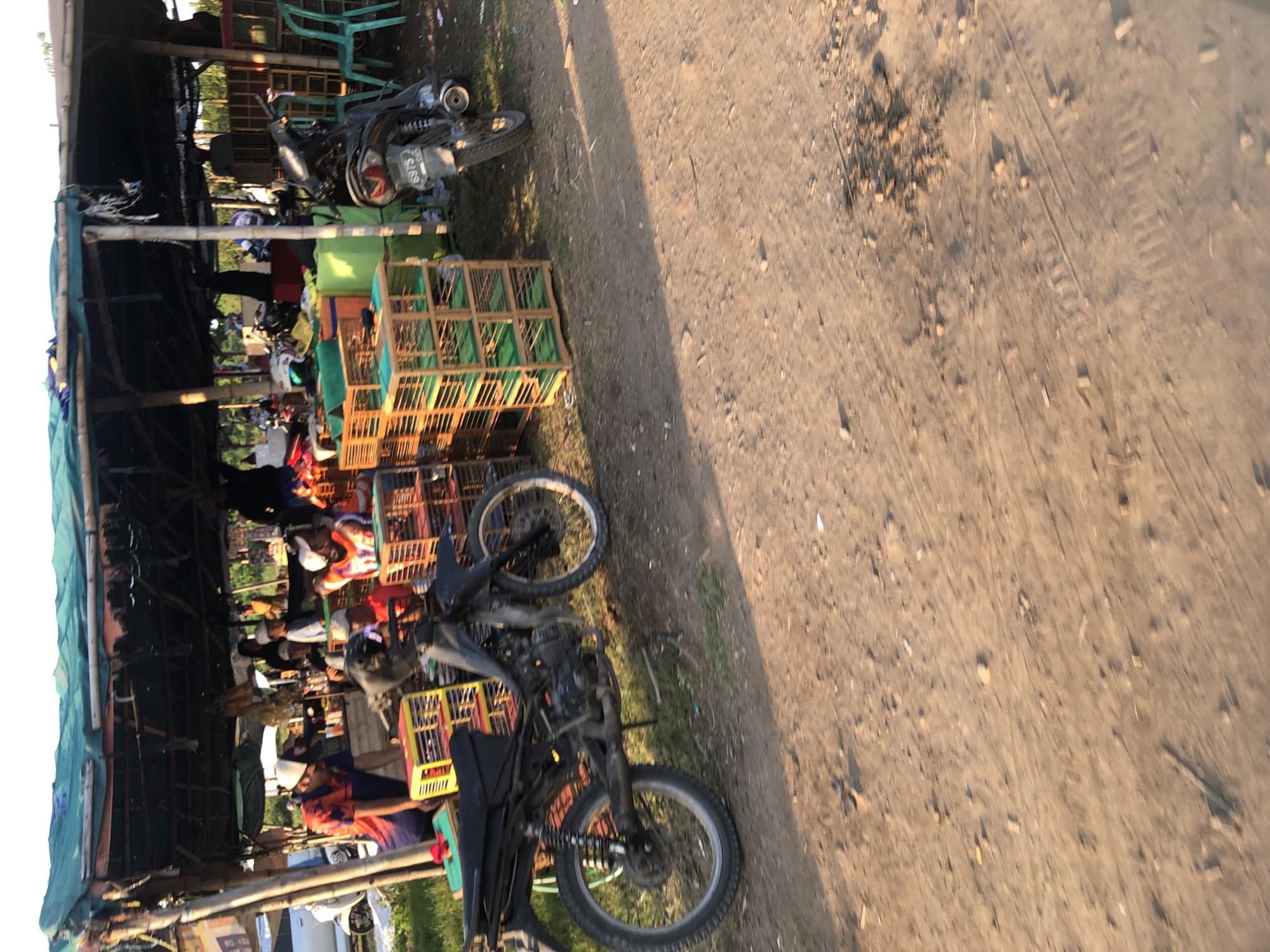 This is a street-level, medium shot taken at an outdoor market in Gempolsari, Indonesia. The scene depicts a stall under a green tarp supported by bamboo poles, with various goods for sale. In the foreground, a black motorcycle is parked, facing left, with its wheels clearly visible. To the left of the motorcycle, a person wearing a colorful shirt and a white cap is leaning over a yellow and red birdcage. Behind them, several other people are partially visible, some wearing caps and engaged in activities around the stall. Stacked wooden birdcages, some with green interiors, are prominent features of the merchandise. In the background, more birdcages and other items are visible under the covered area. To the right, a black motorcycle with a license plate reading "6975" is parked. Green plastic chairs are also visible in the background. The ground is dirt and gravel, indicating an unpaved area, typical of outdoor markets. The overall impression is a bustling market scene with goods and people interacting under a makeshift shelter.