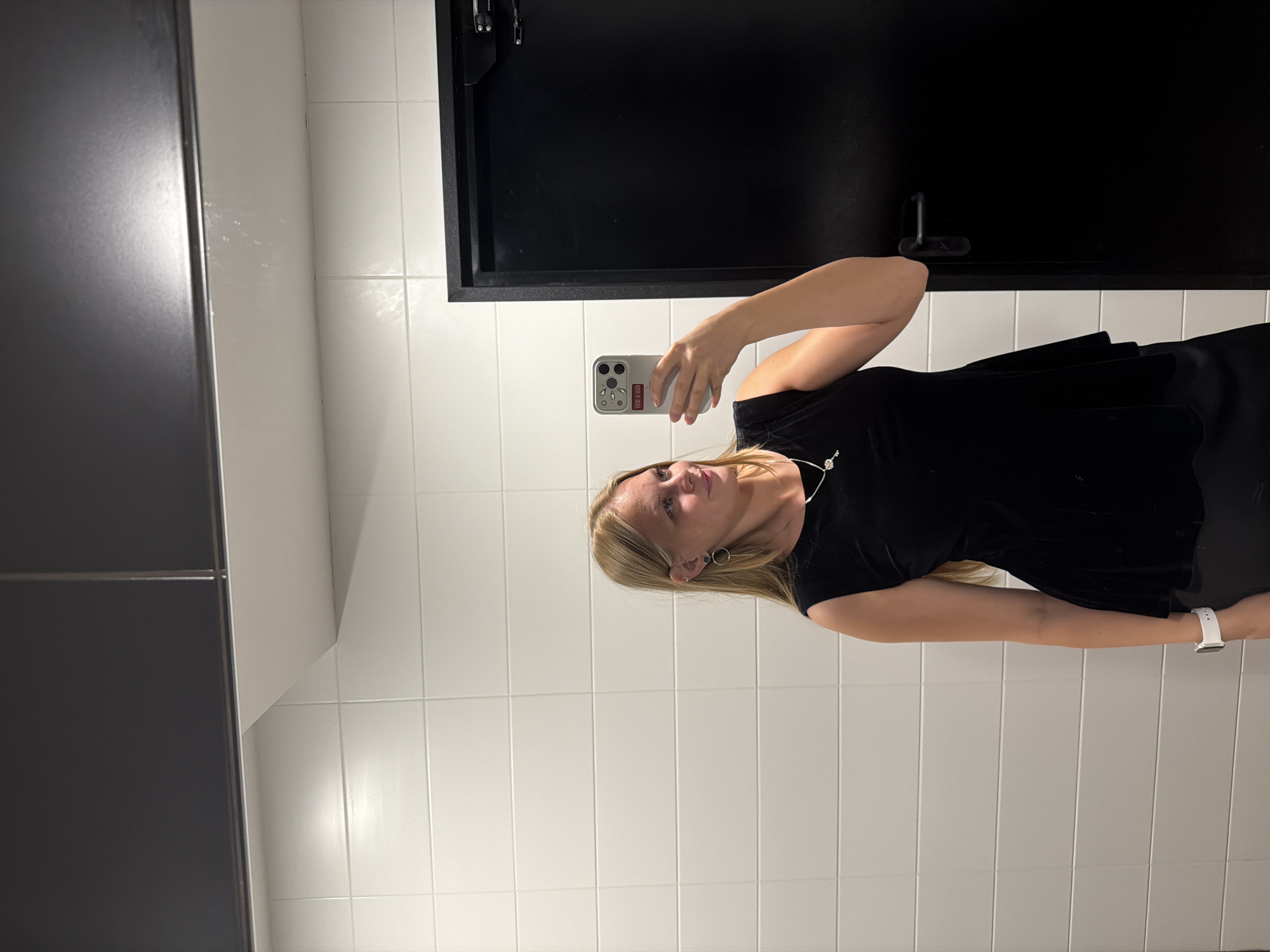The image shows a young woman taking a selfie in what appears to be a public restroom. She has blonde hair and is wearing a black top and a necklace. She is also wearing a white watch on her left wrist. The background is tiled with white square tiles, and there is a black door with a handle. The lighting suggests it is daytime, but there is no information about the weather. The woman's expression is neutral. The image also contains a smartphone with some stickers on the back. There is no visible text in the image.