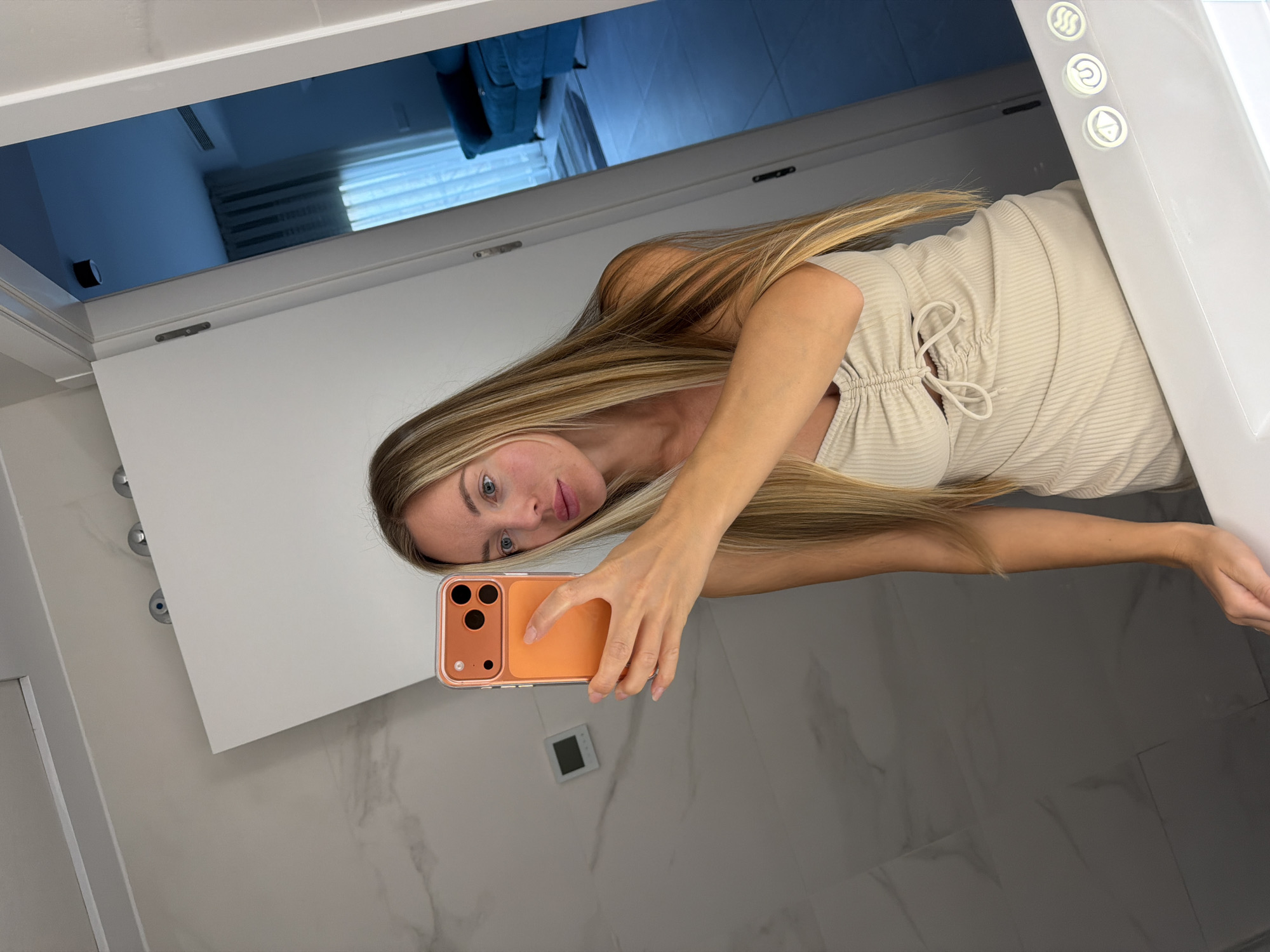 A fair-skinned blonde woman with long blonde hair takes a mirror selfie in what appears to be a modern bathroom. She is wearing a light beige ribbed dress with a drawstring detail at the bust. She holds an orange-cased smartphone in her right hand to capture the reflection. The bathroom features large marbled tiles on the walls and floor, and a white vanity counter. A doorway to the right reveals a glimpse of another room with blue furniture and a blue curtain. The overall impression is one of a clean, well-lit, and contemporary living space, likely in a residential property.