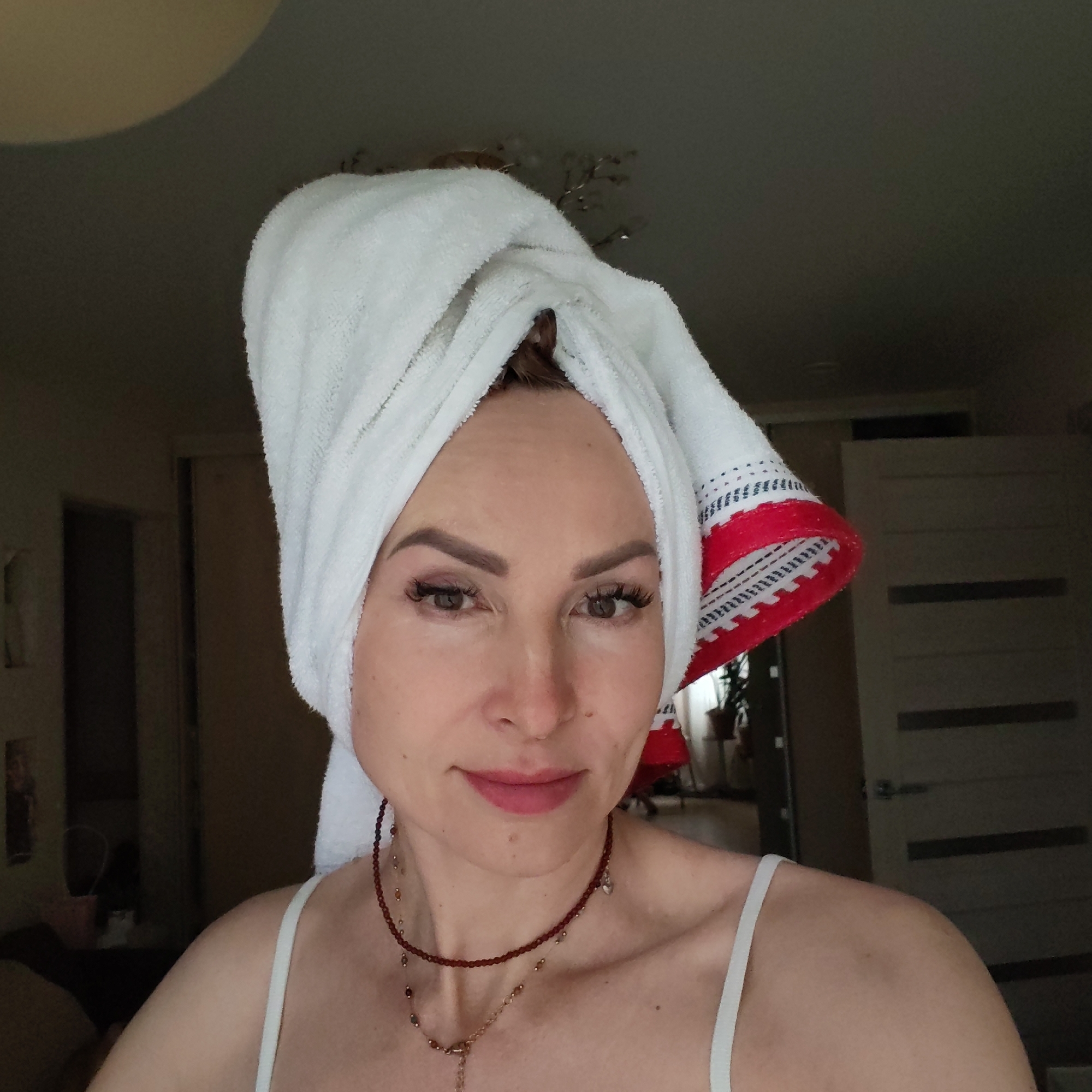 A woman is centered in the frame, wearing a white towel wrapped around her head and a white spaghetti strap top. She has fair skin, dark eyebrows, and what appears to be light pink lipstick. She is also wearing a dark beaded necklace with a pendant. The background shows an interior with open doorways leading to other rooms. A plant is visible through one of the doorways. The overall lighting is somewhat dim. The city of Chaikovsky, Russia, is not visually apparent from the image.