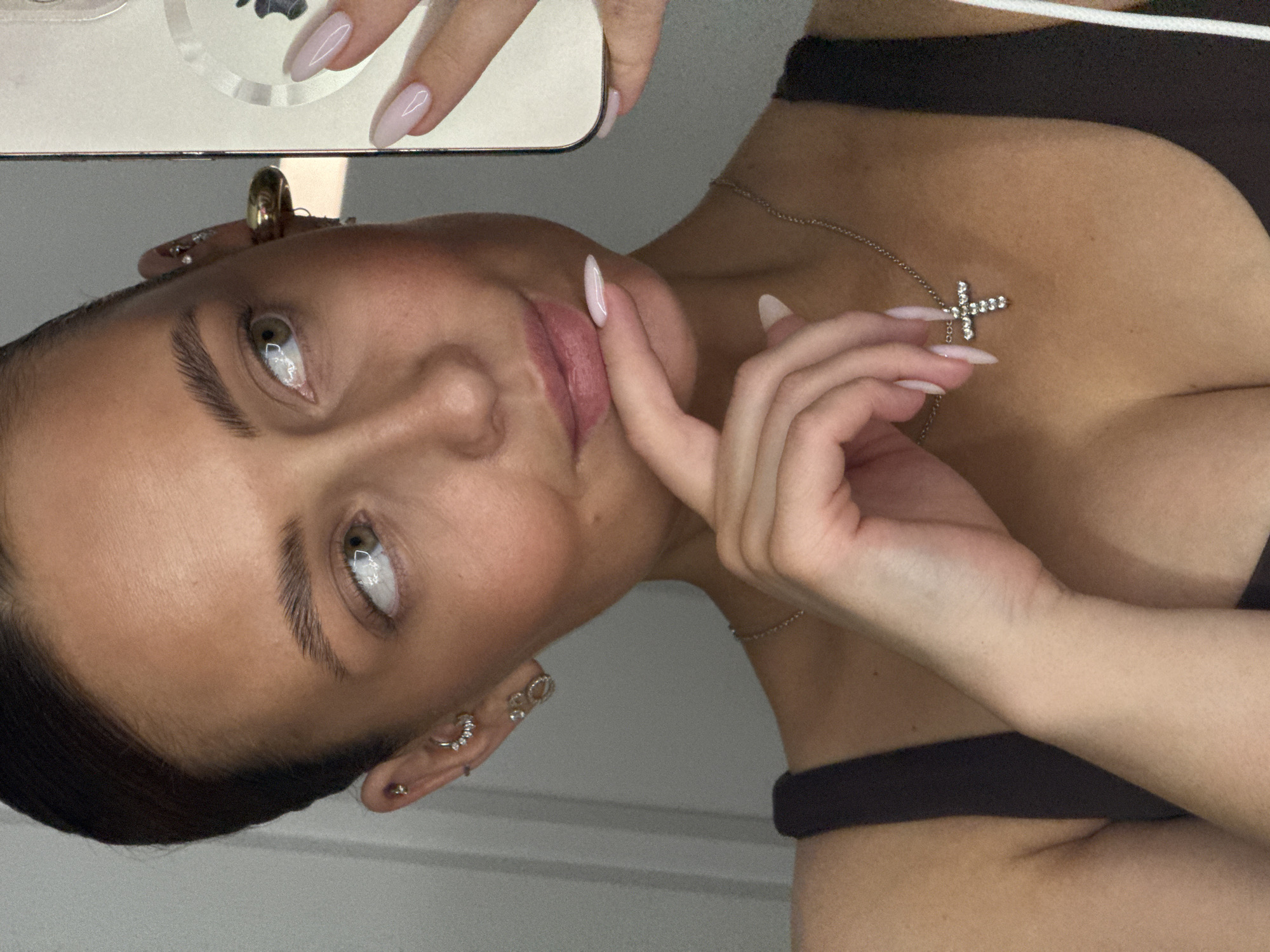 A woman is taking a selfie. She is wearing a brown top and a silver necklace with a cross pendant. Her nails are painted a light pink color. She has multiple piercings in her left ear. The background is blurry and appears to be a white wall. There is no indication of location in the city of Chaikovsky, Russia.