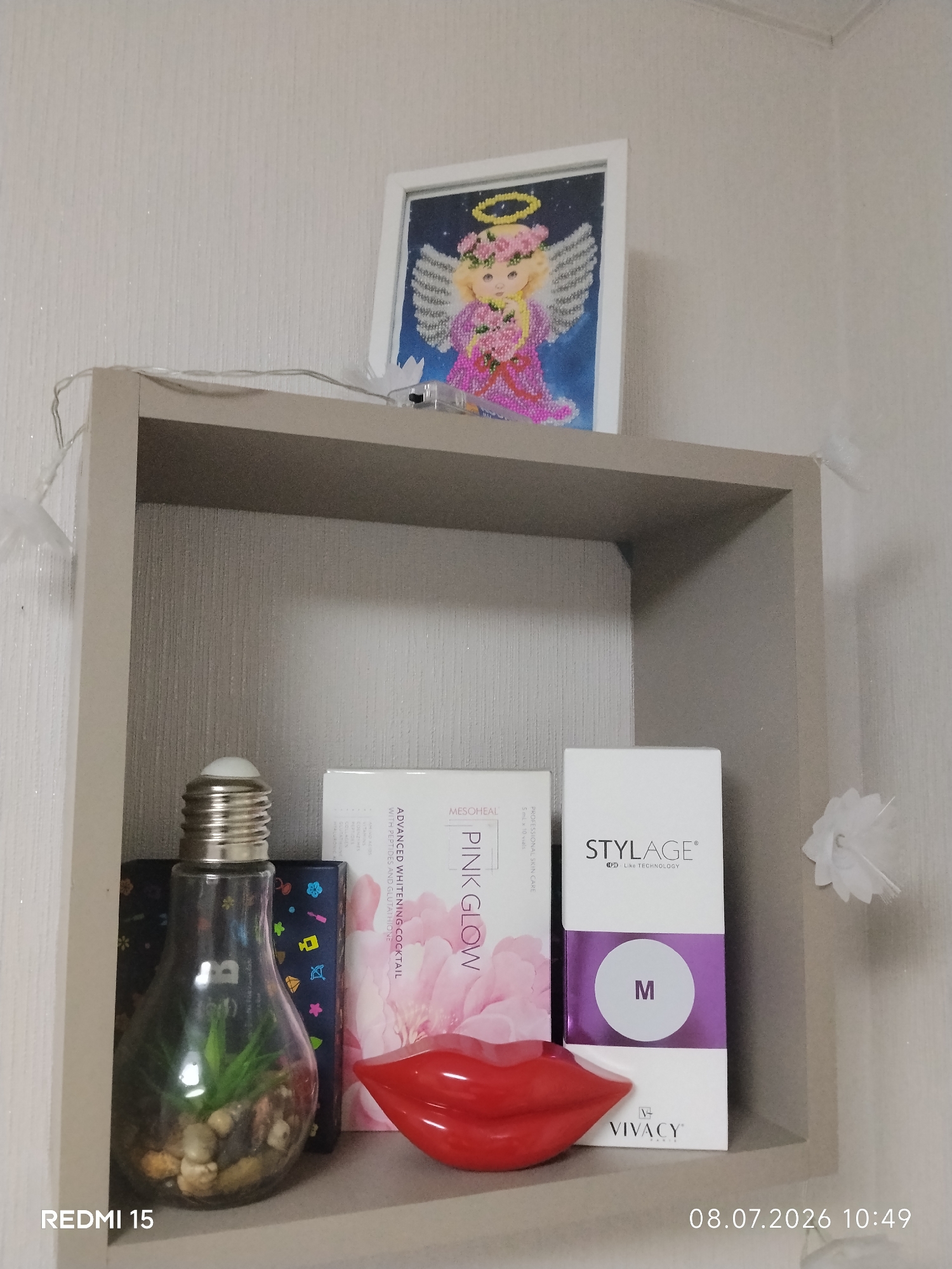 The image shows a shelf with several decorative and cosmetic items. On the top shelf, there is a framed picture of an angel made of beads. Below this, on the lower shelf, there is a glass lightbulb-shaped terrarium with a plant and stones inside. Next to it are two boxes, one labeled "MESOHEAL PINK GLOW" and another labeled "STYLAGE". There is also a red, lip-shaped object. In the background, the wall is textured. The image is a photograph taken indoors. The timestamp "08.07.2026 10:49" and the text "REDMI 15" are visible at the bottom.