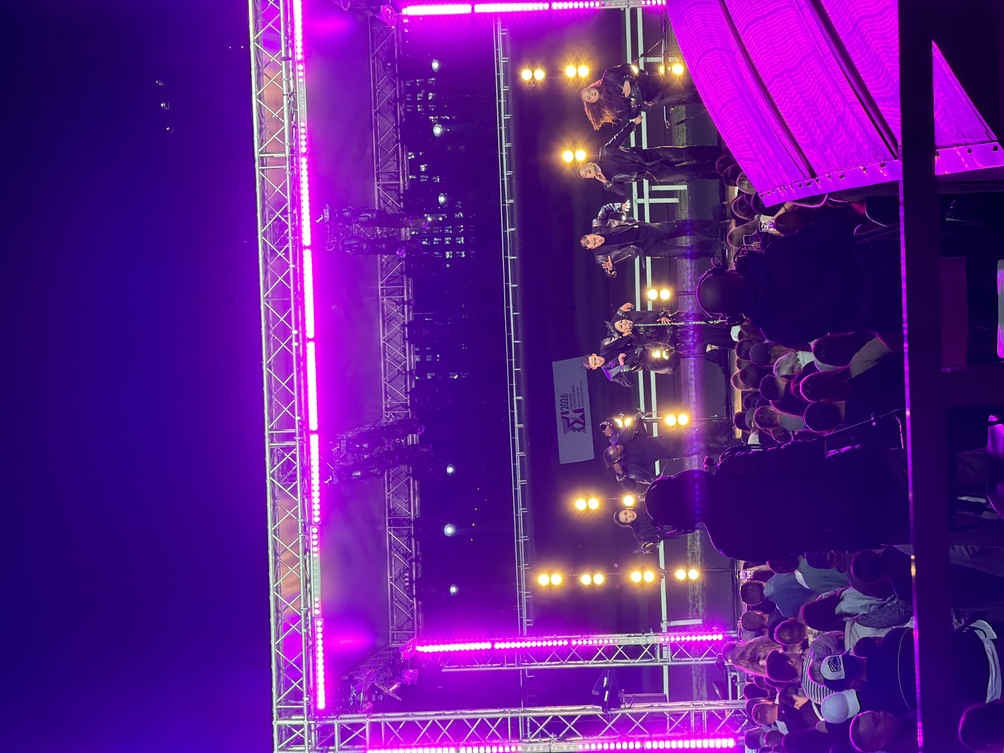 The image depicts an outdoor concert scene at night, illuminated by vibrant purple stage lighting. In the foreground, a crowd of spectators is visible, some holding up their phones to capture the performance. A person in the center foreground is wearing headphones and holding a camera, appearing to be a photographer or videographer.
On the stage, a group of performers is actively engaged in a dance routine. They are dressed in dark, leather-like outfits. Several yellow spotlights are positioned on the stage, creating dramatic lighting effects. Behind the performers, a banner with the text "2026" and some logos is displayed. The stage structure is a metal truss system, with vertical LED light bars contributing to the overall purple illumination. The background suggests an open area, possibly an outdoor venue or park, with a track or railing visible behind the stage. The sky is dark, indicating nighttime.