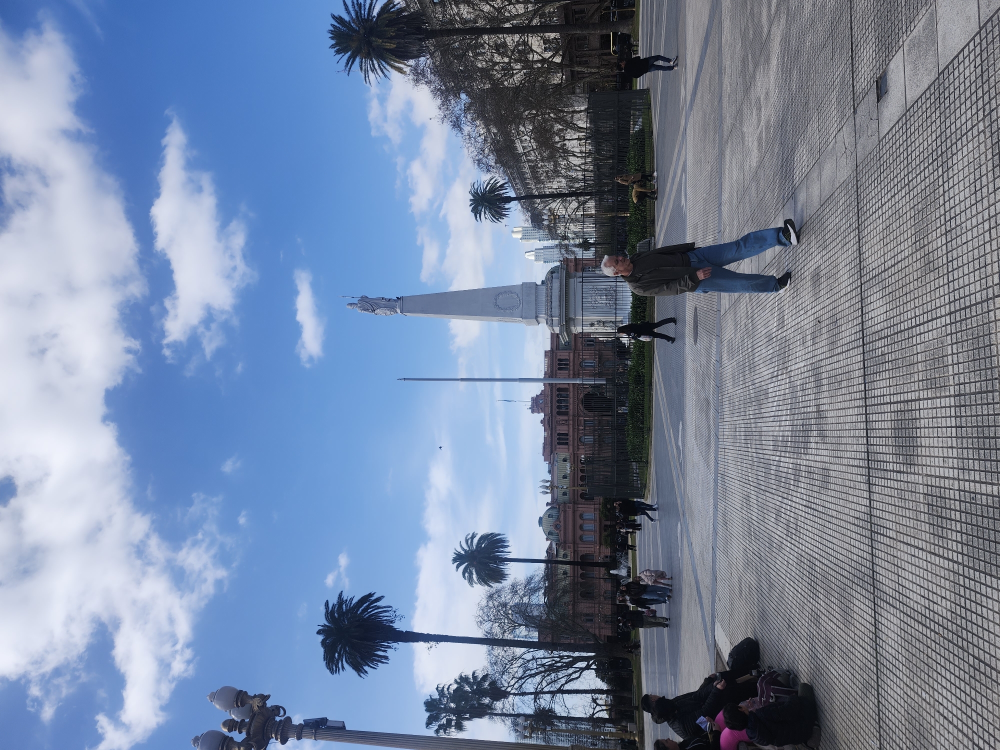 The image captures a daytime view of the Plaza de Mayo in Buenos Aires, Argentina, with the Casa Rosada, the presidential palace, visible in the background. The sky is a bright blue with scattered white clouds. In the foreground, a man in jeans and a jacket walks across a paved square with a grid pattern. Several other people are present in the plaza, some walking and others sitting on the ground or a bench on the left side of the frame. A tall, ornate lamppost stands to the left of the image. In the middle ground, a prominent obelisk-shaped monument topped with a statue stands behind a metal fence. Palm trees and other trees are scattered throughout the scene, with the architecture of the Casa Rosada and other buildings lining the square providing a historical urban backdrop.