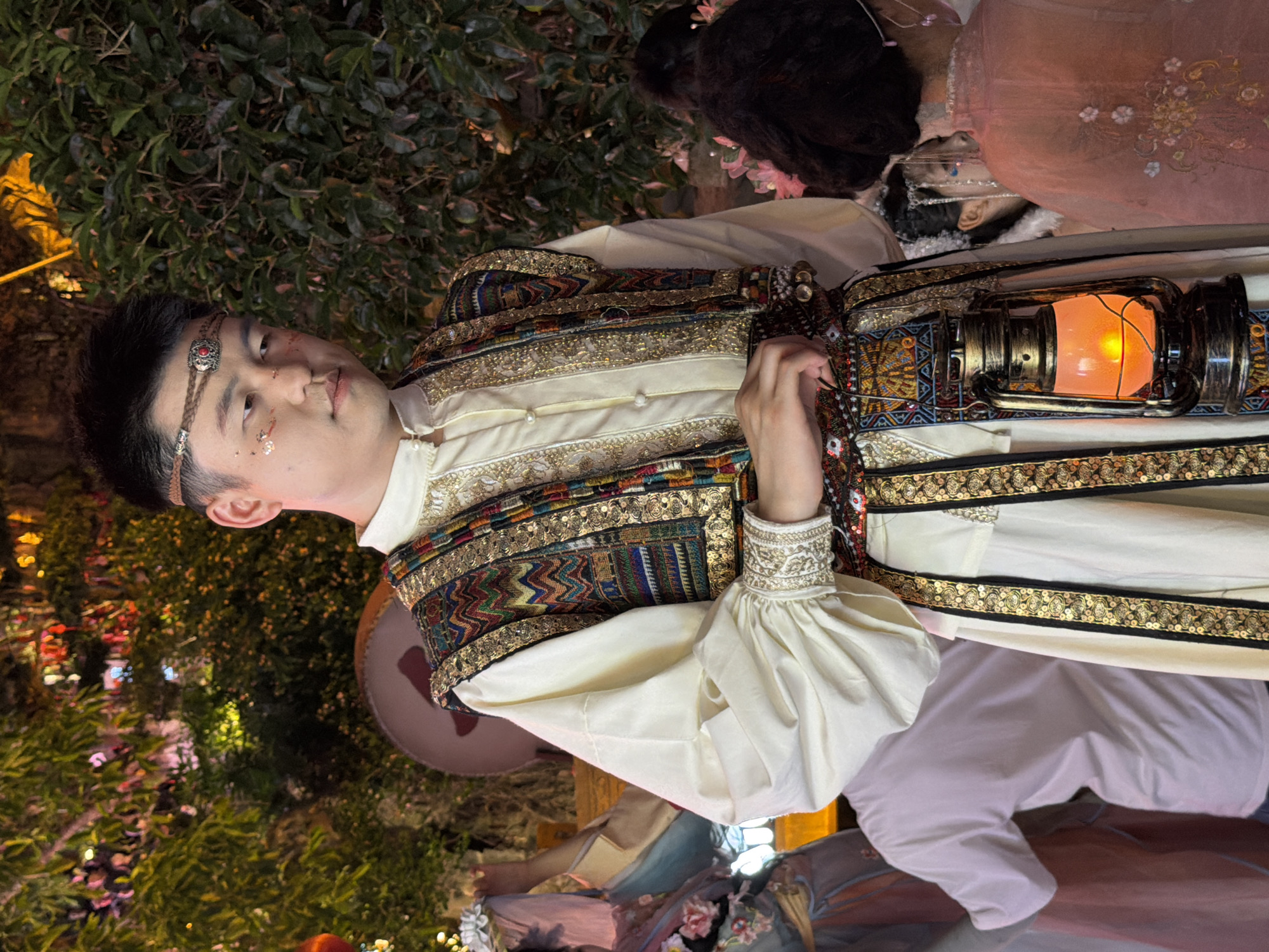 In a dimly lit, outdoor setting, likely a festival or cultural event in Yuzhong District, China, a young East Asian male stands center frame. He is adorned in traditional attire, featuring a cream-colored tunic with voluminous sleeves and a richly embroidered, multi-colored vest. The vest is embellished with gold trim and various patterns. A decorative, beaded belt cinches his waist, from which hangs an antique-style oil lantern, its flame casting a warm orange glow. His hair is short and dark, and he wears a braided headband with a central red embellishment. Subtle facial makeup, including what appears to be glitter or colored powder, is applied to his cheekbones.
Behind him, to the left, another person is partially visible, wearing a light pink t-shirt. Further in the background, lush green foliage is illuminated by decorative lights, suggesting an evening event. To the right of the central figure, the back of a female is visible, wearing a pink, sheer garment with embroidered floral designs. Another person, likely a female, is seen in the background between the central male and the woman in pink, also wearing elaborate traditional clothing and headwear. The overall atmosphere is festive and culturally rich.
