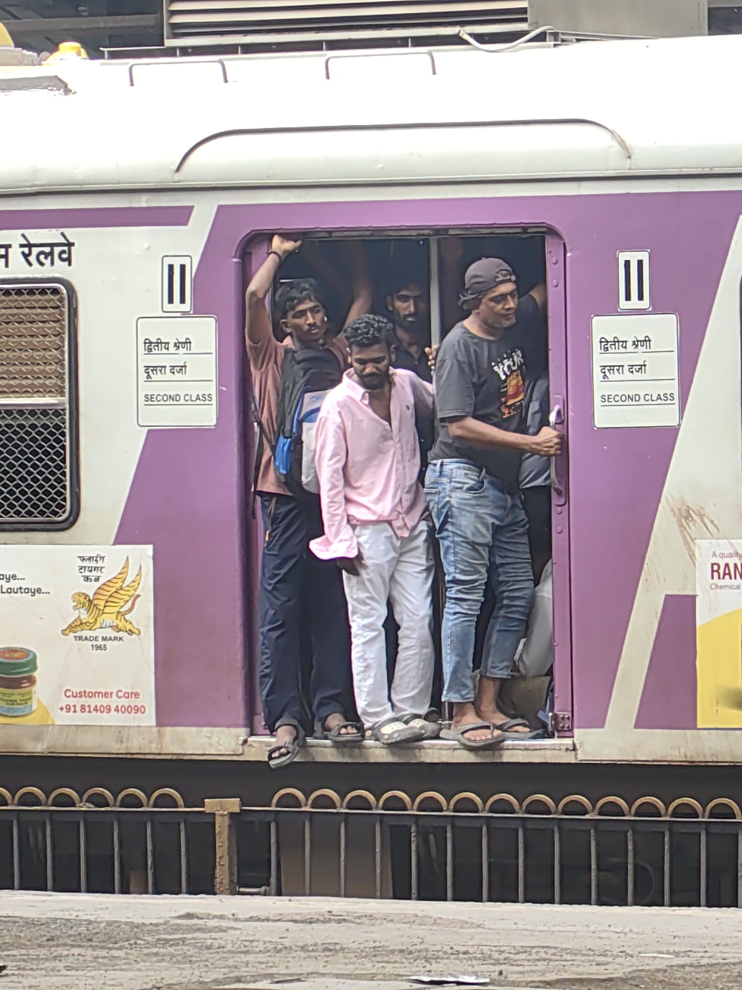 A group of men are crammed into the doorway and interior of a purple and white commuter train car in Mira Bhayandar, India. The men are standing very close together, with several of them leaning out of the open doorway. One man in a pink shirt is visible in the center, wearing white pants. Another man in a dark t-shirt and jeans is holding onto the train door handle. The train car has "SECOND CLASS" signage in both Hindi and English. A yellow and red advertisement featuring a winged tiger is affixed to the side of the train car. The scene appears to depict a crowded public transportation environment.