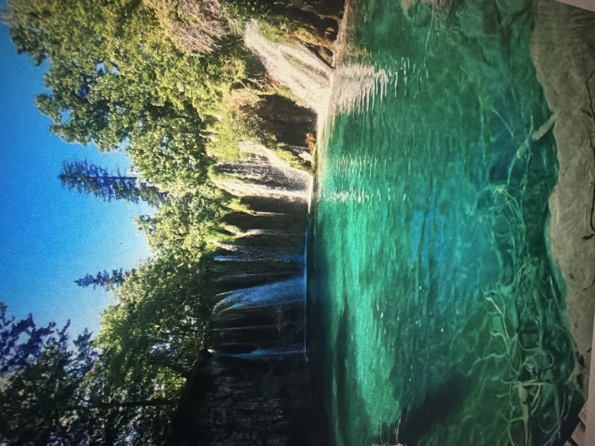 This image depicts a vibrant, turquoise pool of water with visible submerged roots and rocks, leading to a series of cascading waterfalls. Lush green trees and foliage surround the waterfalls, forming a natural amphitheater. The sky above is a clear, bright blue. There are no people or man-made structures visible in this natural landscape. The scene is tranquil and picturesque, showcasing the beauty of a pristine natural environment. The image does not provide any information or visual cues that place it specifically in Baan Mae Nam, Thailand.