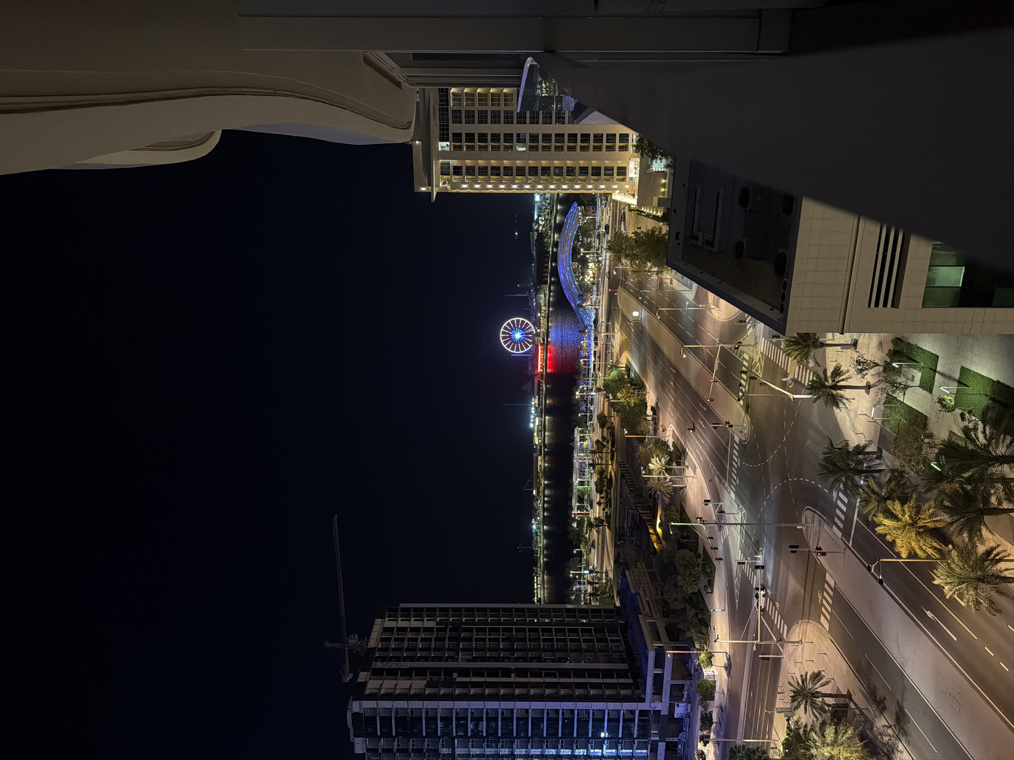 The image captures a nighttime, elevated perspective of an illuminated urban waterfront from a balcony in Unknown, Unknown. In the distance, a large Ferris wheel glows with red, blue, and white lights across a body of water, with other illuminated buildings and structures lining the far shore. A prominent arched bridge, lit in blue, spans part of the waterway closer to the foreground. To the left, a tall, multi-story concrete building under construction features a large crane extending from its top, with some of its lower sections showing blue illumination. To the right, a completed, light-colored multi-story building with numerous warmly lit windows partially frames the scene, with a beige balcony railing and wall obscuring the extreme right foreground. Below, a network of multi-lane asphalt roads with white markings, intersections, and traffic lights is visible, bordered by numerous illuminated palm trees and landscaped green spaces. A few vehicles, including cars and a motorcycle, are sparsely distributed on the roads. The sky is dark and clear.