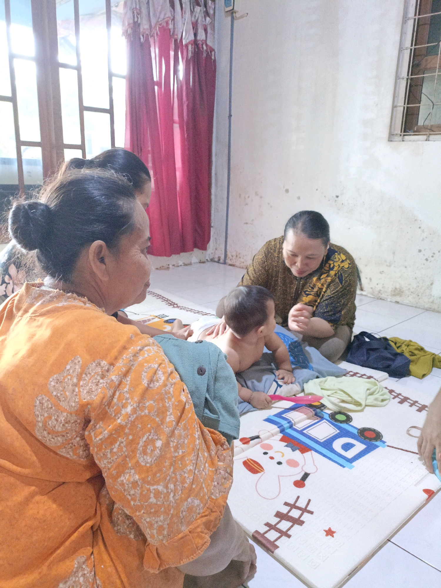 In the city of Jenu, Indonesia, an indoor scene unfolds with a woman in an orange patterned garment in the foreground, facing away from the camera. Behind her, another woman in a dark patterned top is seated on a mat with a young child. The child, appearing to be a toddler, is partially dressed and facing the seated woman, who is looking down at the child. Various clothing items, including a yellow garment, are spread on the mat near the seated woman and child. The mat itself has a colorful design featuring a cartoon bus and a rabbit figure. The background shows a window with sunlight streaming through pink curtains, and a textured wall with some discoloration. The flooring appears to be tiled.