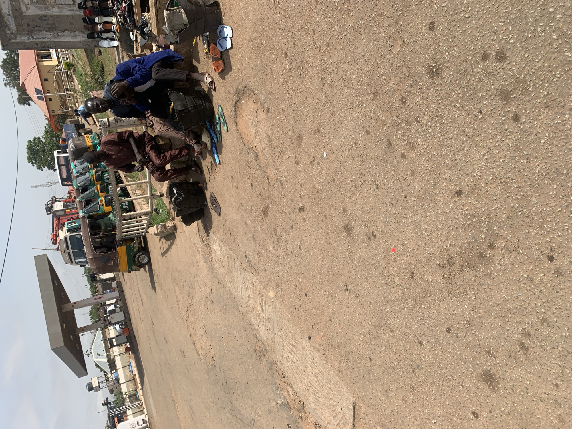 The scene is on a street in Gura Top, Nigeria. Two men are crouched next to each other on the side of the road, both appearing to be fixing something. The man on the left wears a brown shirt, and the man on the right wears a blue shirt. Several three-wheeled vehicles, some green and some yellow, are parked on the side of the road. There is a gas station in the background. Several pairs of sandals are scattered near the men. The road is paved, and the sky is overcast.
