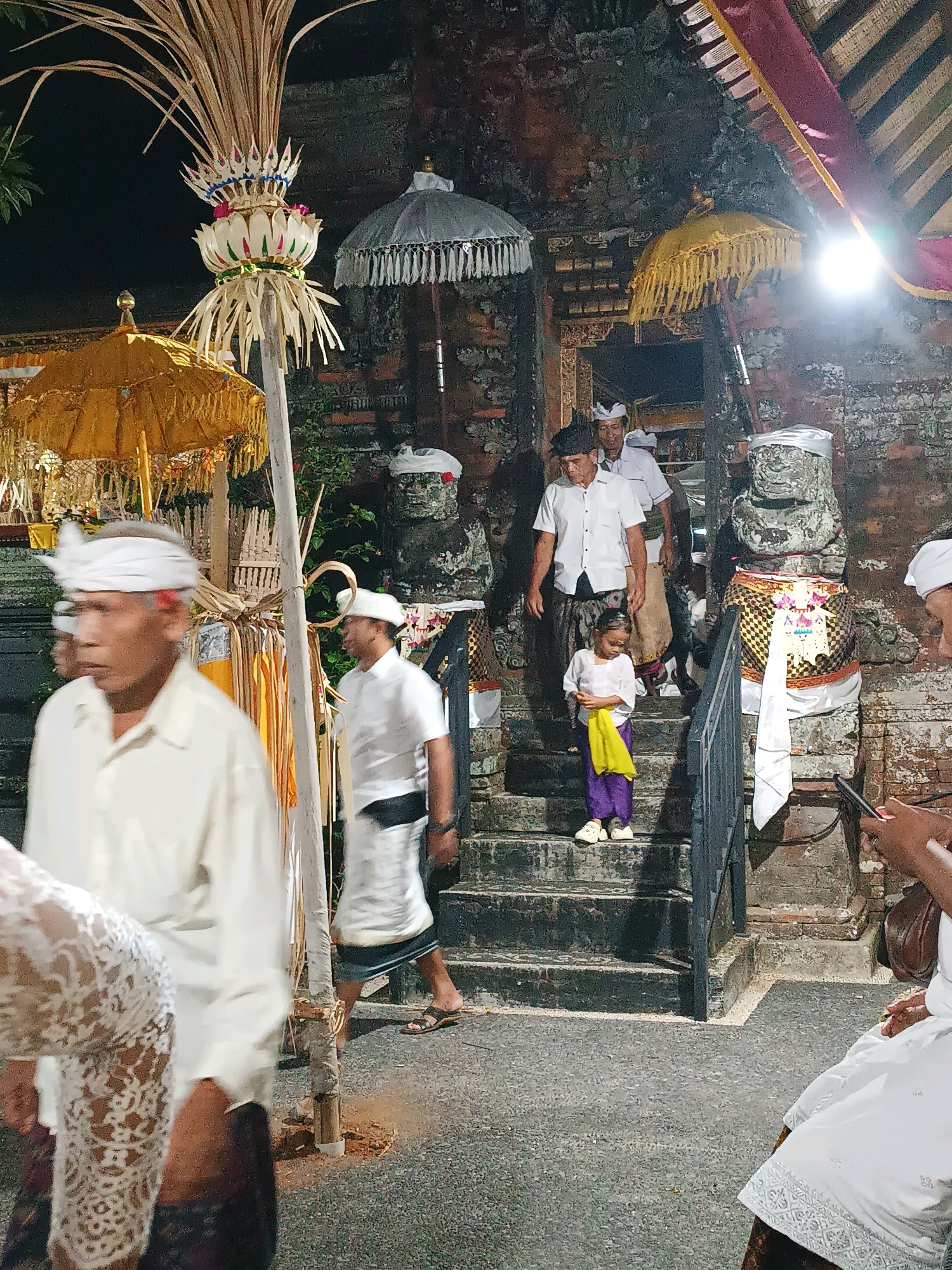 The image depicts a nighttime scene at what appears to be a temple or cultural site in Kediri, Indonesia. Several people, dressed in traditional attire, are visible. In the foreground, a man in a cream-colored shirt and white head covering is partially visible on the left, looking away from the camera. Behind him, a man in a white shirt and white head covering walks down a stone staircase. To the right of the staircase, a young girl in a white top and purple pants, holding a yellow cloth, stands on the stairs. Further up the stairs, two men in white shirts and sarongs are walking.
The architecture suggests a traditional Balinese or Javanese temple with stone structures and ornate decorations. Decorative umbrellas and large, intricate floral arrangements on poles are present. The ground is paved with asphalt, and the lighting indicates artificial illumination, possibly from lamps or ambient light from nearby buildings. The overall atmosphere suggests a religious ceremony or cultural event.