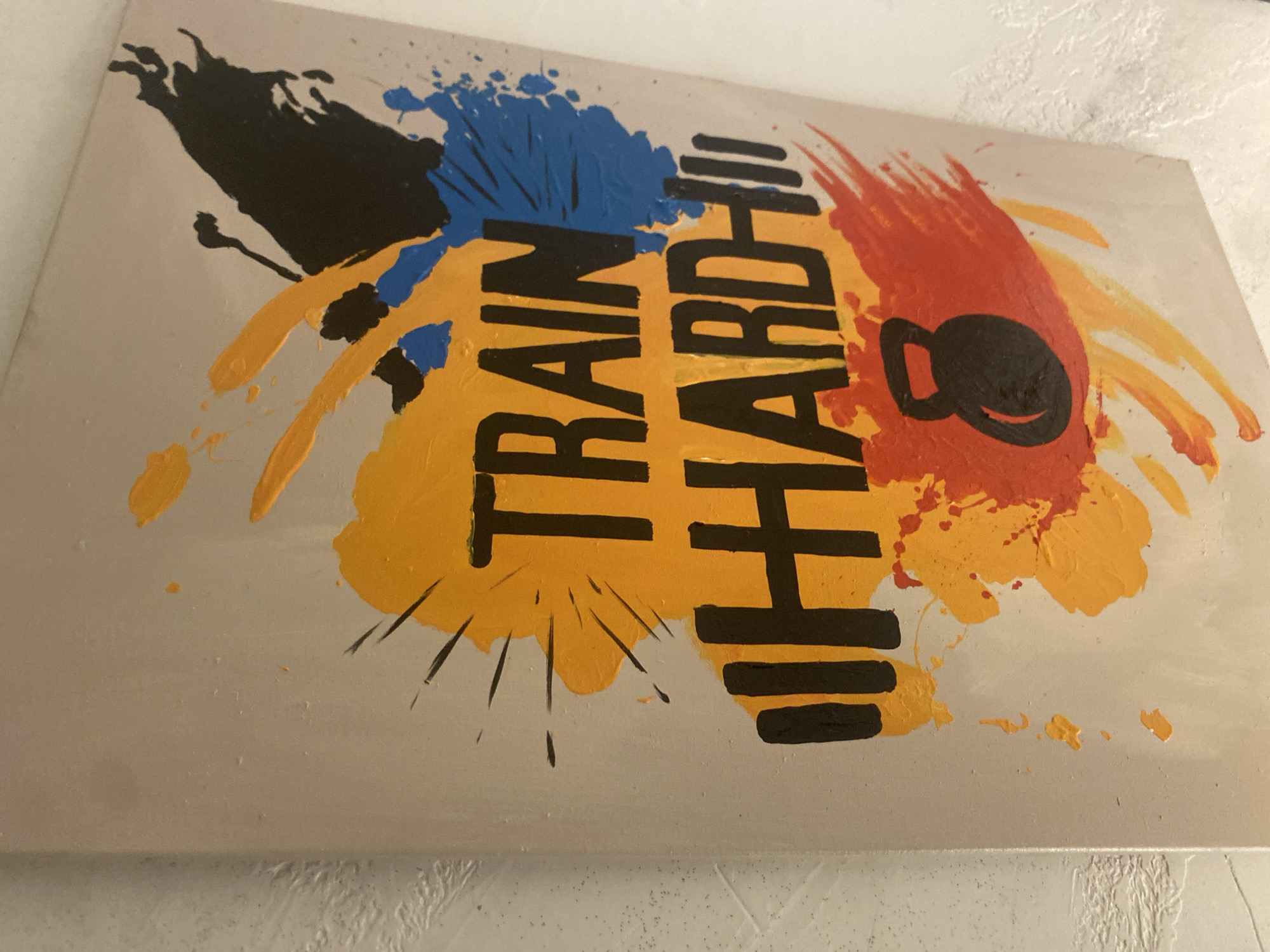 This is a close-up shot of a painting on a wall. The painting features bold, black lettering that spells out "TRAIN HARD" with a dumbbell icon to the right of the word "HARD". The lettering is set against a background of colorful paint splatters in shades of yellow, blue, and red. The overall style is energetic and abstract. The painting appears to be a motivational piece, likely for a gym or fitness-related space. The image does not depict any specific location in Barden Yana, Nigeria, nor does it show any people or actions.