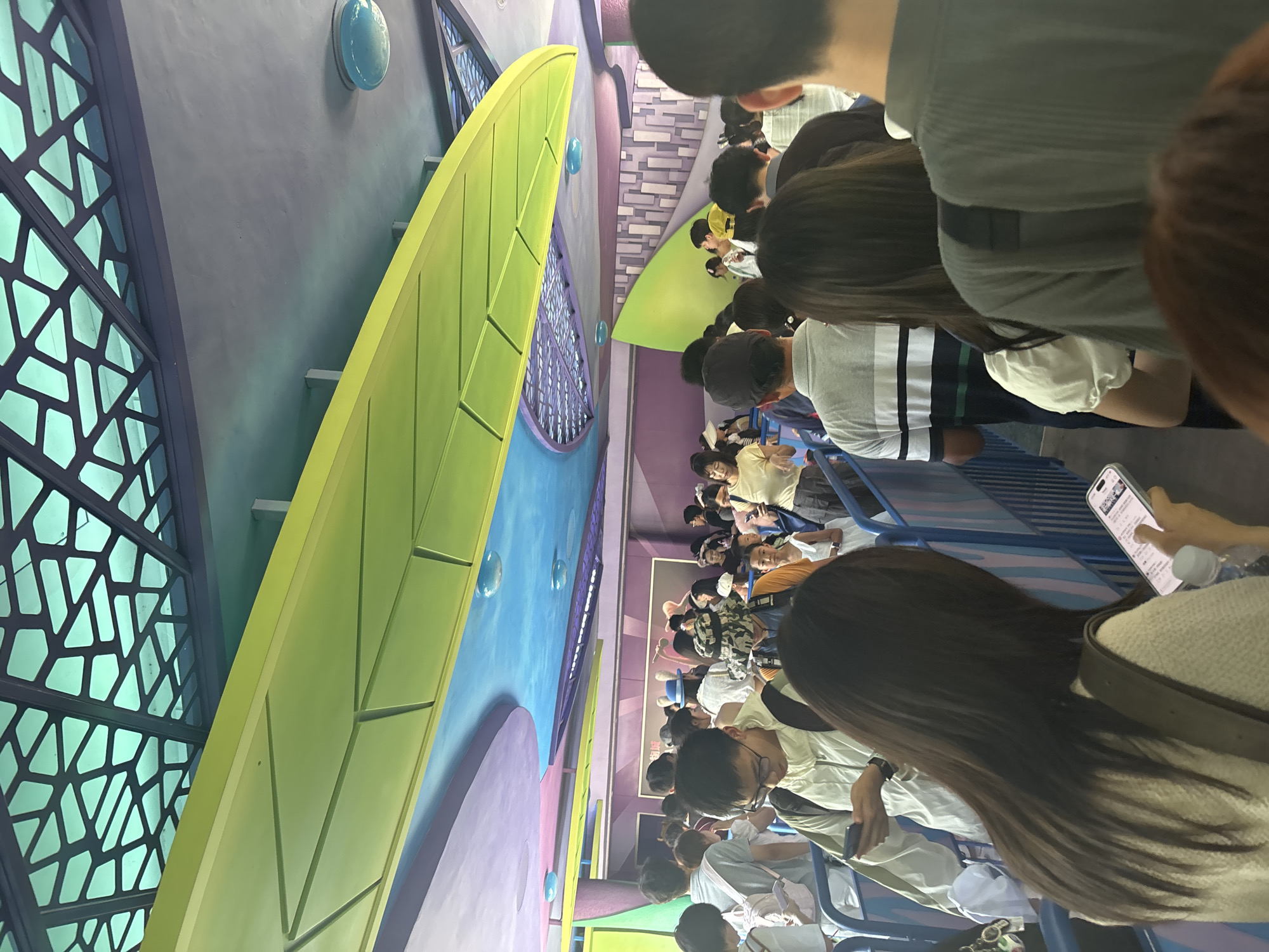 In Pudong, China, a crowd of people is queuing in a brightly colored indoor environment. The ceiling features abstract, organic shapes in lime green and purple, with a patterned translucent green structure overhead. Blue circular lights are embedded in the ceiling. The queue barriers are blue and white. People are dressed in casual attire, and many are looking at their mobile phones or talking to each other. Some individuals are holding backpacks. The overall atmosphere suggests a theme park or entertainment venue.