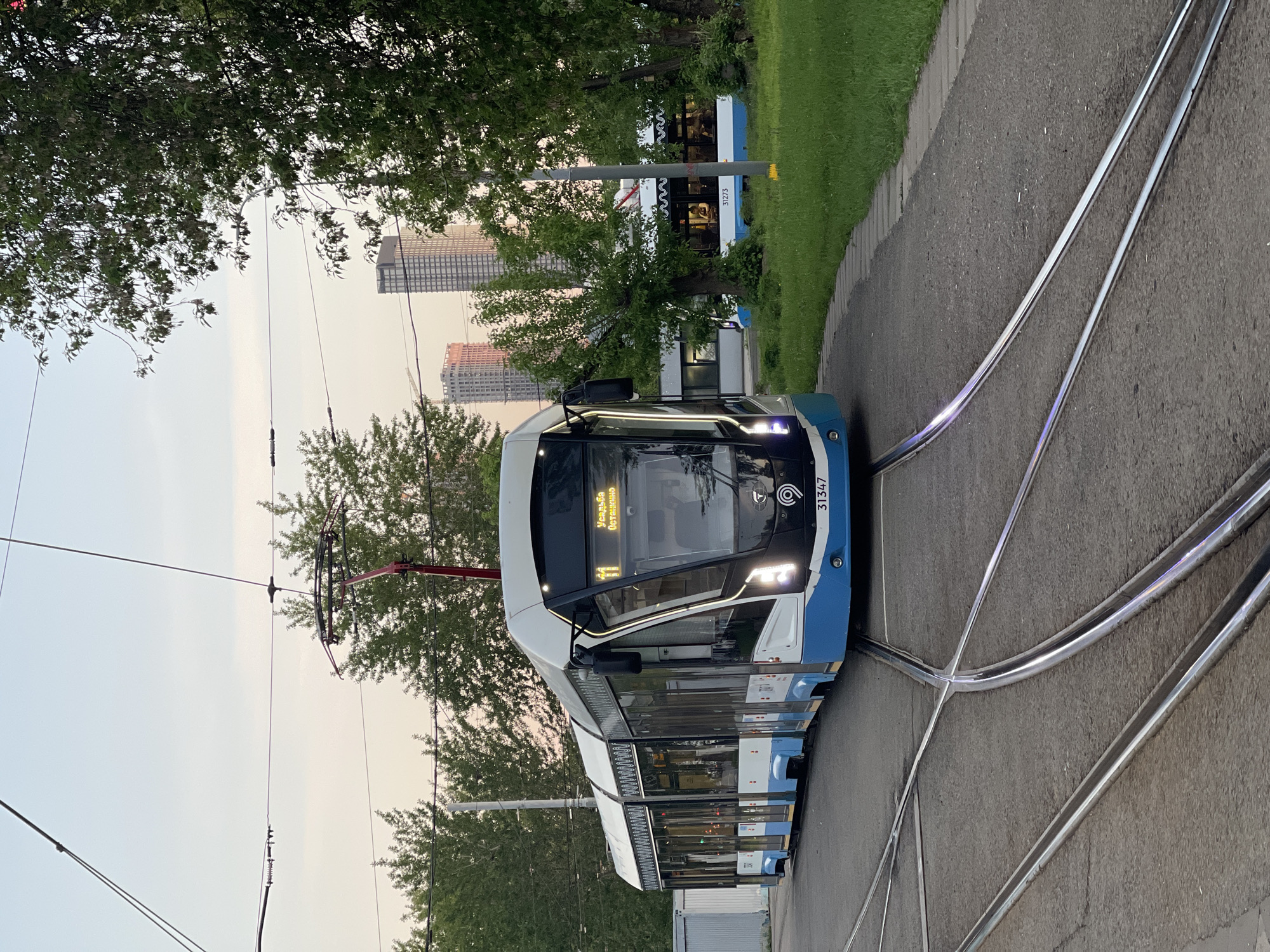 A tram is moving on a track. The tram is white and blue. The sign on the tram indicates it is going to Ulitsa Ostanino. Tall buildings are in the background. Trees are on the side of the street.