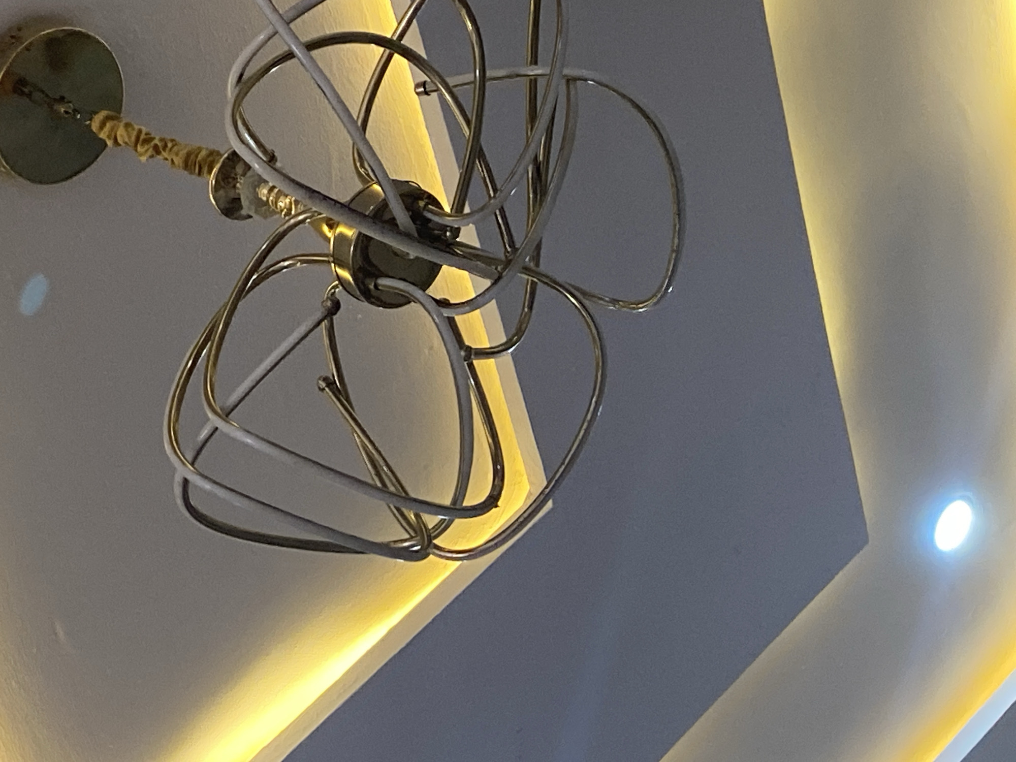 A close-up, angled shot from below shows a modern, ornate ceiling light fixture with a tangled, wire design. The fixture is composed of gold-colored metal rods that curve and intersect to form a somewhat abstract shape. Some rods are also covered in white. The fixture is attached to the ceiling by a short, covered pole connected to a circular base. The surrounding environment consists of a ceiling with recessed lighting and clean, sharp angles. The lighting gives the ceiling a soft glow, with cooler gray tones dominating the shadowed areas and warm white tones highlighting the architectural lines. The exact location is not identifiable.