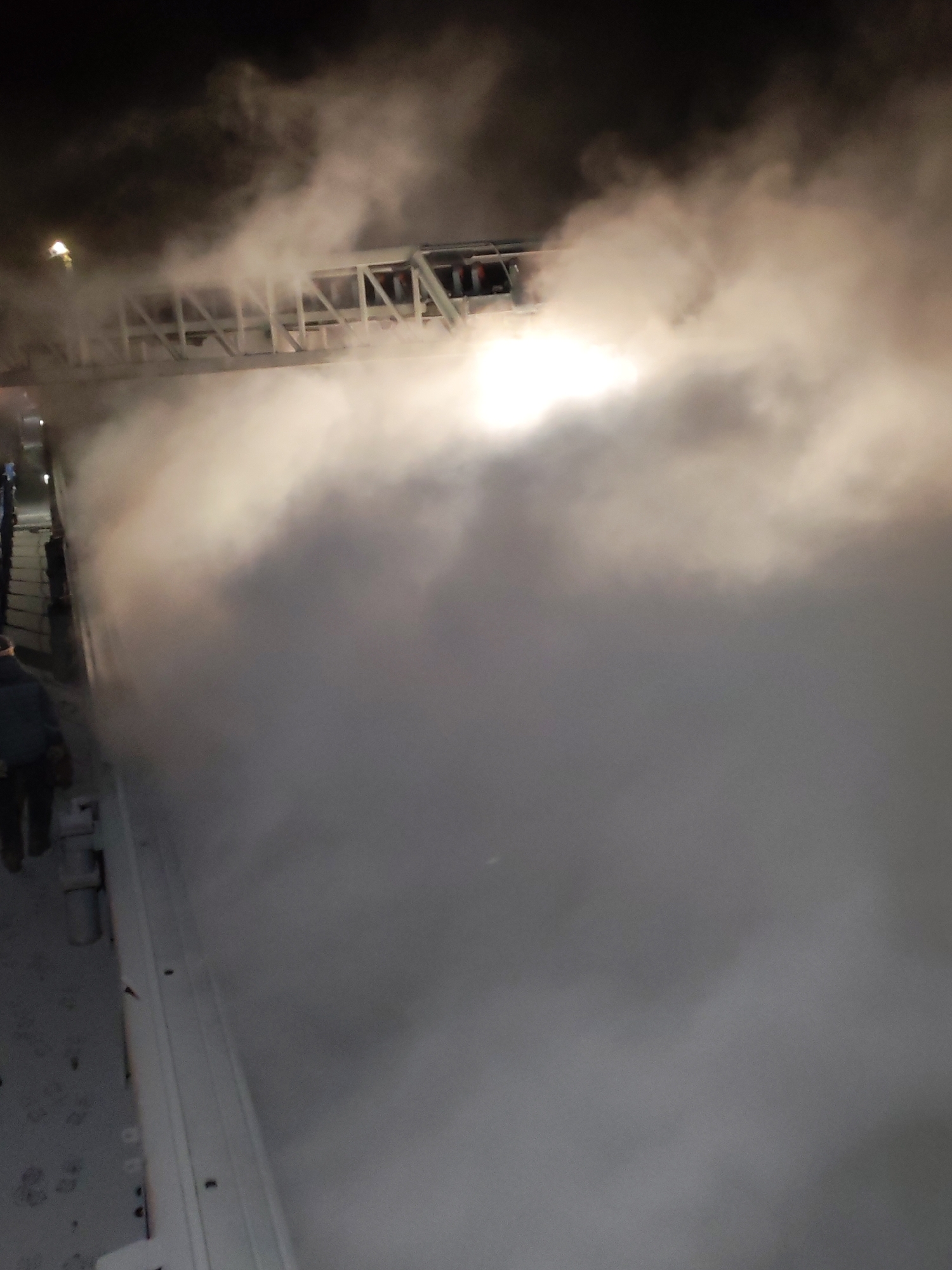 The photograph depicts a scene at night, heavily obscured by fog or smoke. A white structure, possibly a bridge or scaffolding, is partially visible through the fog. A bright light source illuminates the fog from behind. On the left side, a person is walking along a surface, possibly a sidewalk or platform. The surrounding environment is dark and appears to be outside. Given the information provided, it is not possible to confirm the precise location as Shcheleiki, Russia, as the image itself does not contain indicators of the city.