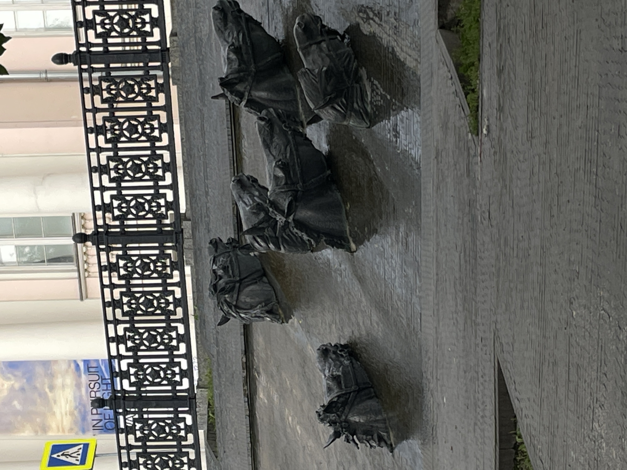 The image captures a portion of the "Four Horsemen of the Apocalypse" sculpture by Zurab Tsereteli, located on a ramp leading up to the Victory Bridge in Moscow, Russia. The sculpture consists of several bronze horse heads emerging from a wet, textured surface, possibly concrete or stone, that slopes upwards. The horse heads are detailed, with visible manes and bridles, and appear to be partially submerged in water or wetness. In the background, a black ornamental metal fence with repeating geometric patterns is visible. Above the fence, a building with light-colored walls and windows can be seen, along with a portion of a large banner displaying text that reads "IN PURSUIT OF LIGHT". A yellow triangular pedestrian crossing sign with a black silhouette of a person walking is also present on the left side of the image. The overall impression is one of a public art installation in an urban setting, with the wet surfaces suggesting recent rain or a water feature.