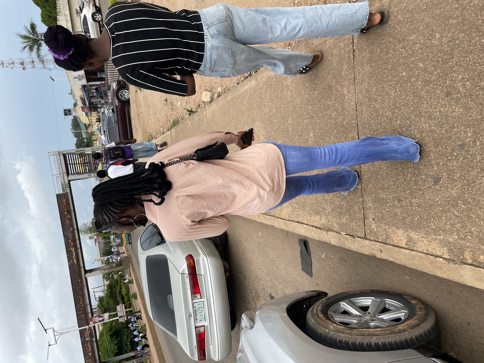 Two women are walking on a sidewalk in Jos, Nigeria, in the afternoon. The woman on the left is wearing a light pink long-sleeved shirt and blue flared jeans. She has black braided hair and is carrying a black purse and a phone. The woman on the right is wearing a black and white striped long-sleeved shirt and light blue wide-leg jeans. She has her hair in a bun with a purple accessory and is wearing sandals. They are walking away from the camera on a concrete sidewalk. To their left, there is a road with several cars, including a silver Toyota Camry. In the background, there are buildings, trees, a traffic light, and a radio tower. The sky is overcast.