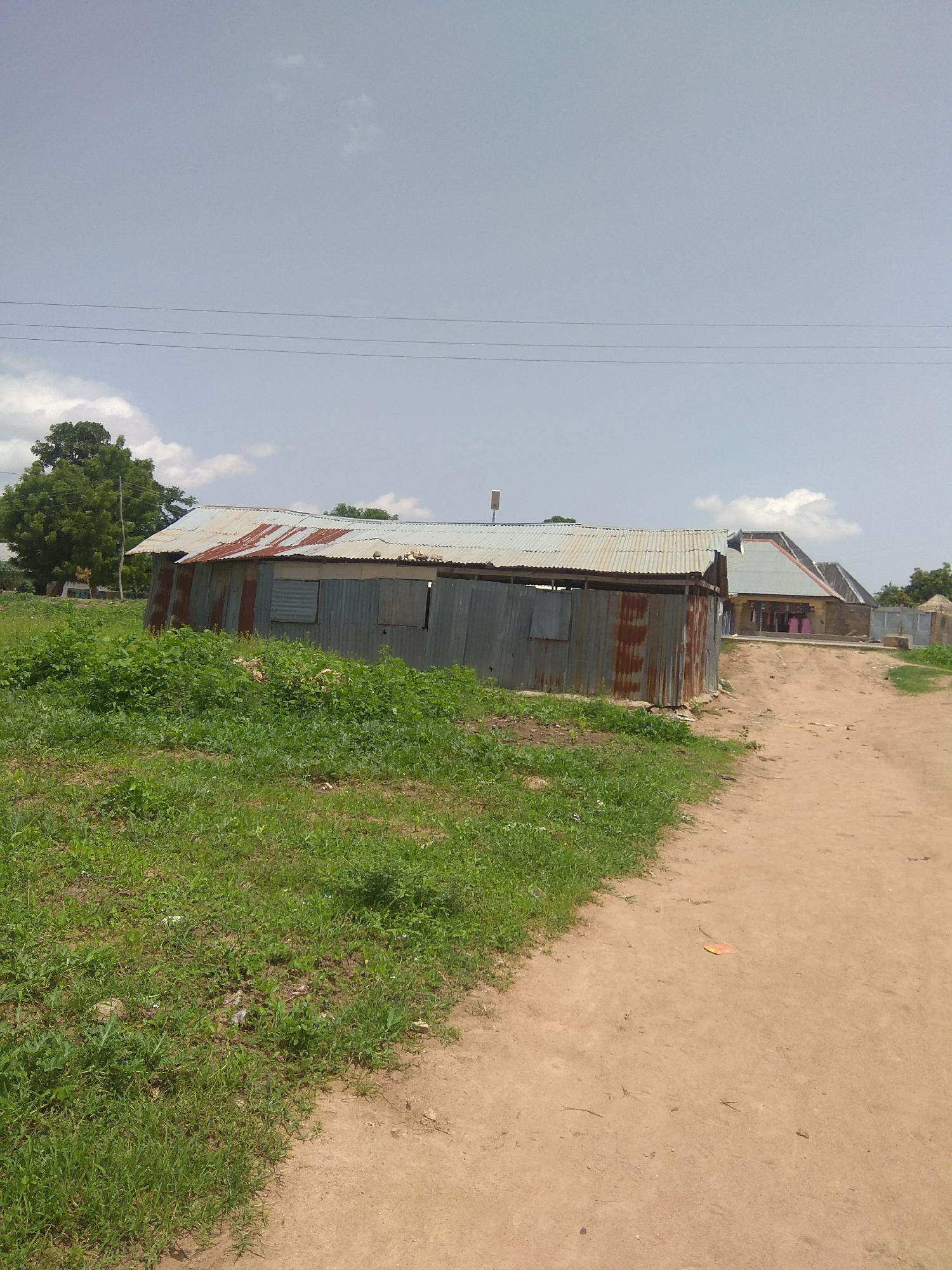 The image depicts an outdoor scene with a dirt road leading towards several buildings in Jalingo, Nigeria. The foreground features a patch of green grass and weeds on the left side, with a rough, sandy path dominating the right. In the mid-ground, a long, low-lying building constructed with corrugated metal walls and roofing, showing signs of rust and age, is visible. This structure has at least two openings that appear to be windows or doorways. Further down the road, a second building with a more complex, angled roof made of corrugated metal can be seen, with a fence in front of it and a hint of other structures beyond. The background includes a large, green tree on the far left, and a clear sky with scattered clouds. Power lines are visible stretching across the sky. The overall impression is of a rural or semi-urban setting, characterized by simple, functional architecture and a natural landscape. There are no people or animals visible in the scene.