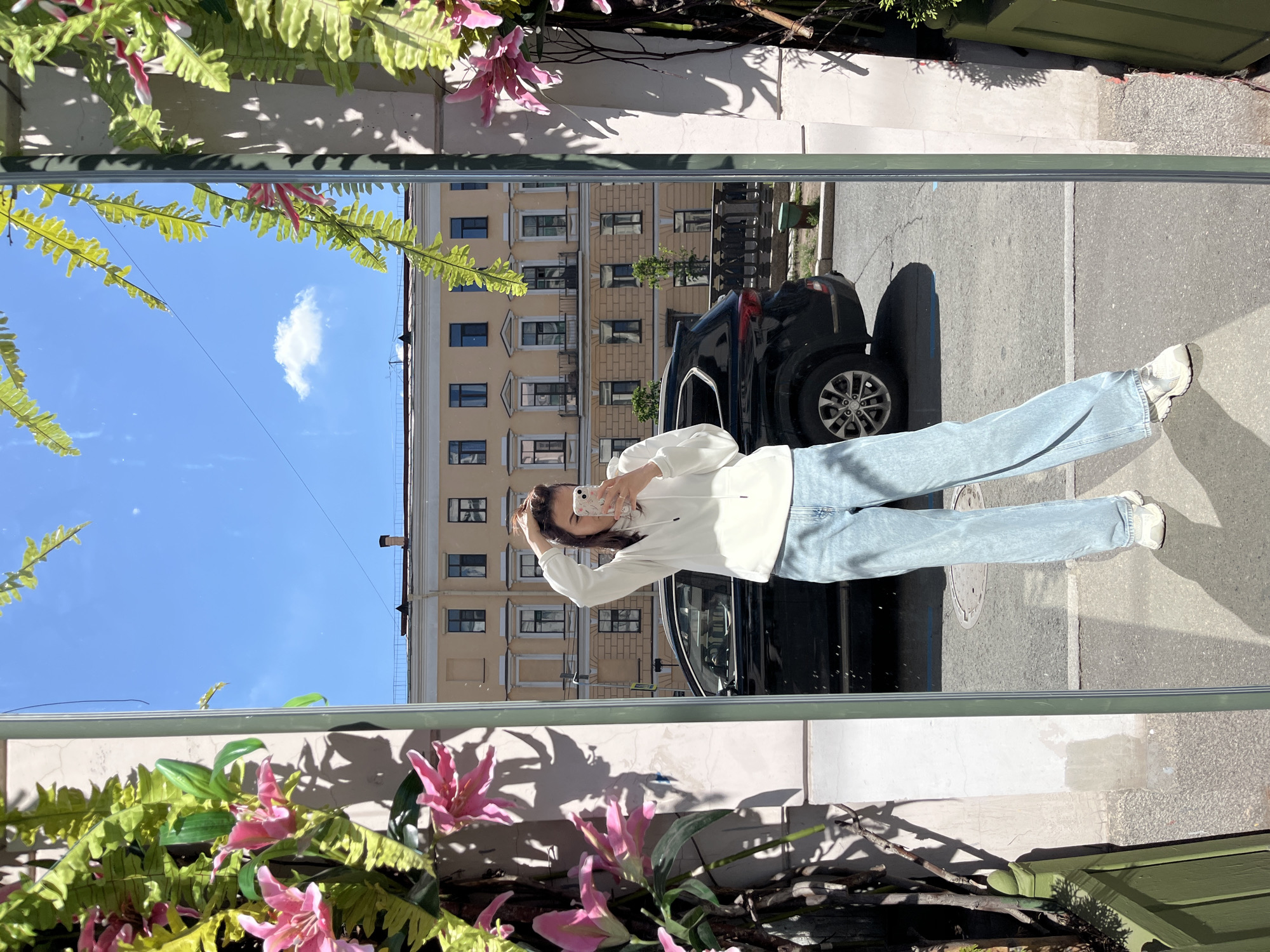 A young woman with dark hair pulled back is reflected in a large, full-length mirror, holding a smartphone and taking a selfie. She is wearing a white hooded sweatshirt and light blue wide-leg jeans, with white sneakers. Her left hand is raised, touching her hair. The mirror is framed by an abundance of artificial greenery and pink lilies, which cast shadows on the wall behind her. The reflection shows a street scene in Saint Petersburg, Russia, with a parked black SUV, a building with yellow facades and numerous windows, and a bright blue sky with a few clouds. The street has a cobblestone and asphalt surface. The overall setting suggests a sunny day in an urban environment.